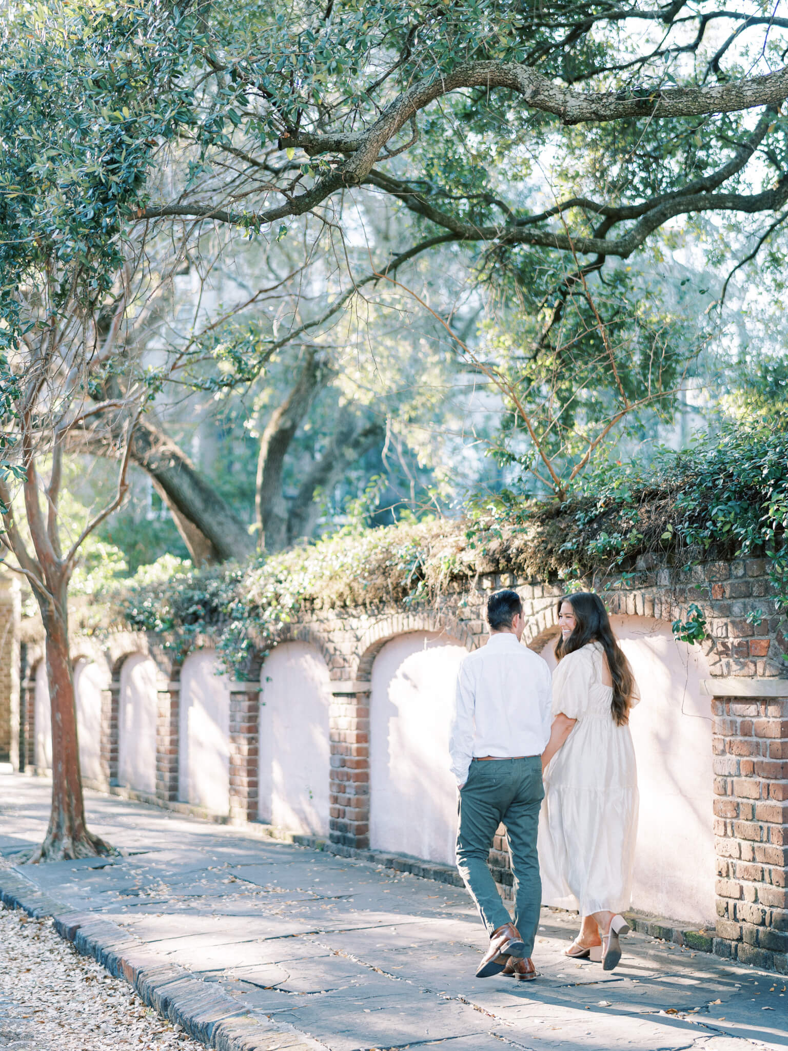 An engaged couple walking away with linked arms on Chalmer's Street with an old wall, greenery and trees surrounding them.