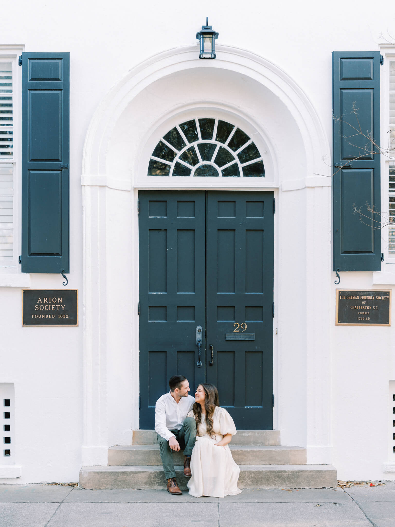 A man and woman smiling at each other while sitting on the stoop of a white building on Chalmer's Street for their Charleston engagement photos.