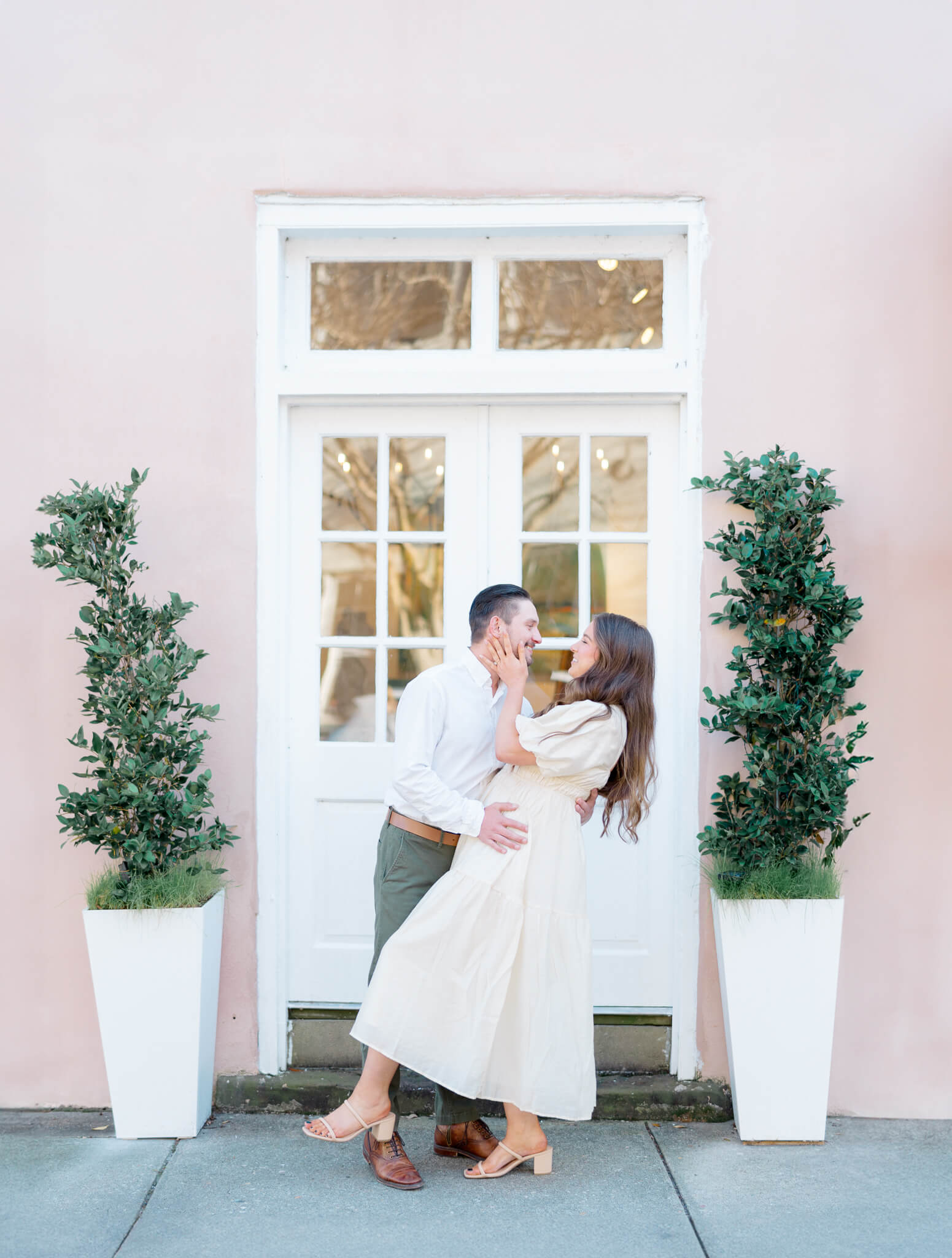 A man dipping a woman back to kiss her in front of a white door and pink building with two shrubs.