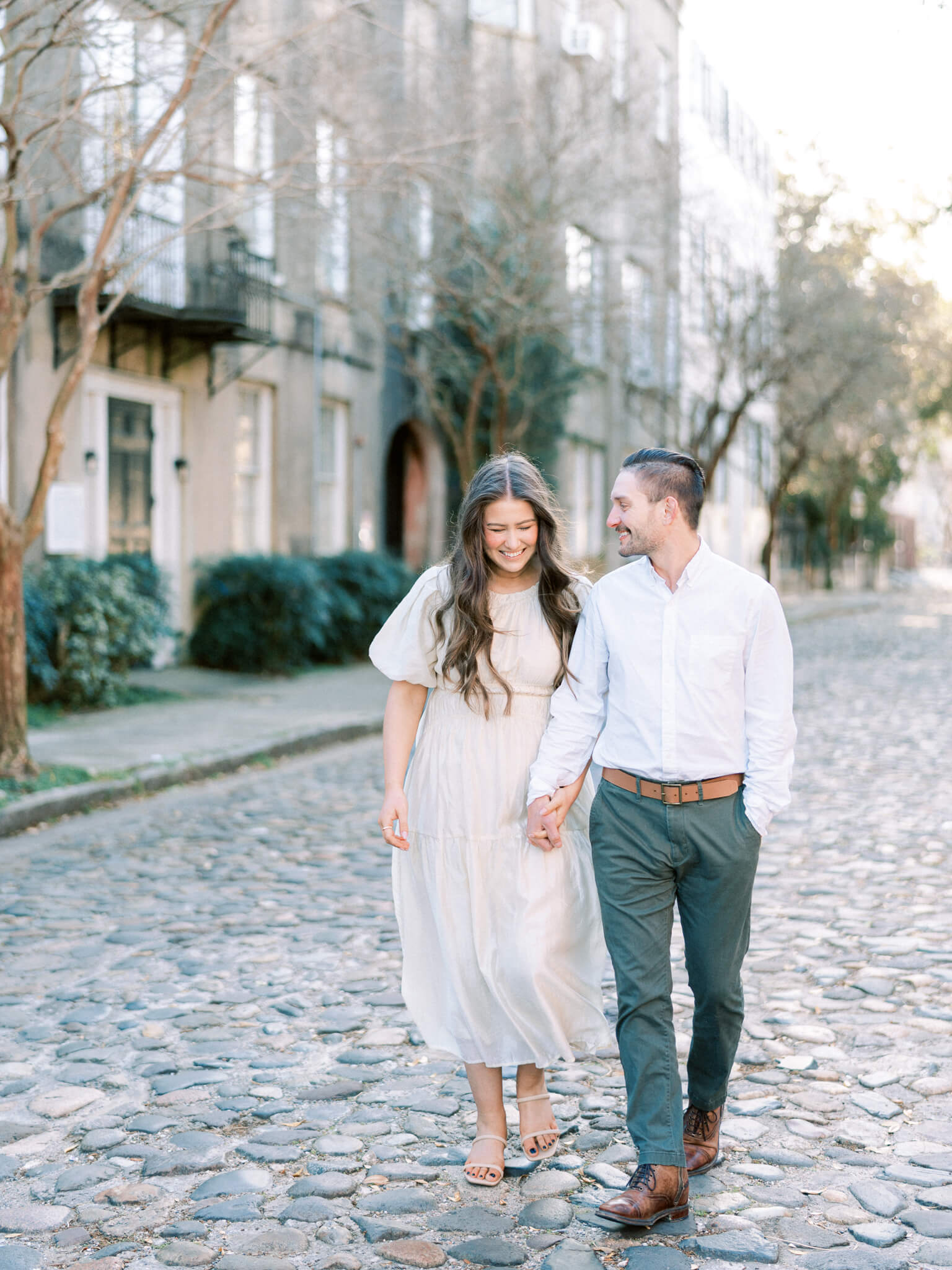 A man in green pants and a white shirt walking hand in hand with a woman in a cream dress on Chalmer's Street in Charleston.