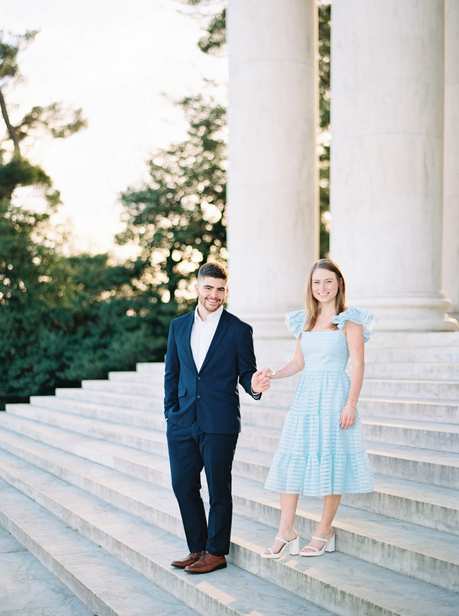 An engaged couple walking down the steps of the Jefferson Memorial with the sun rising and marble columns in the background