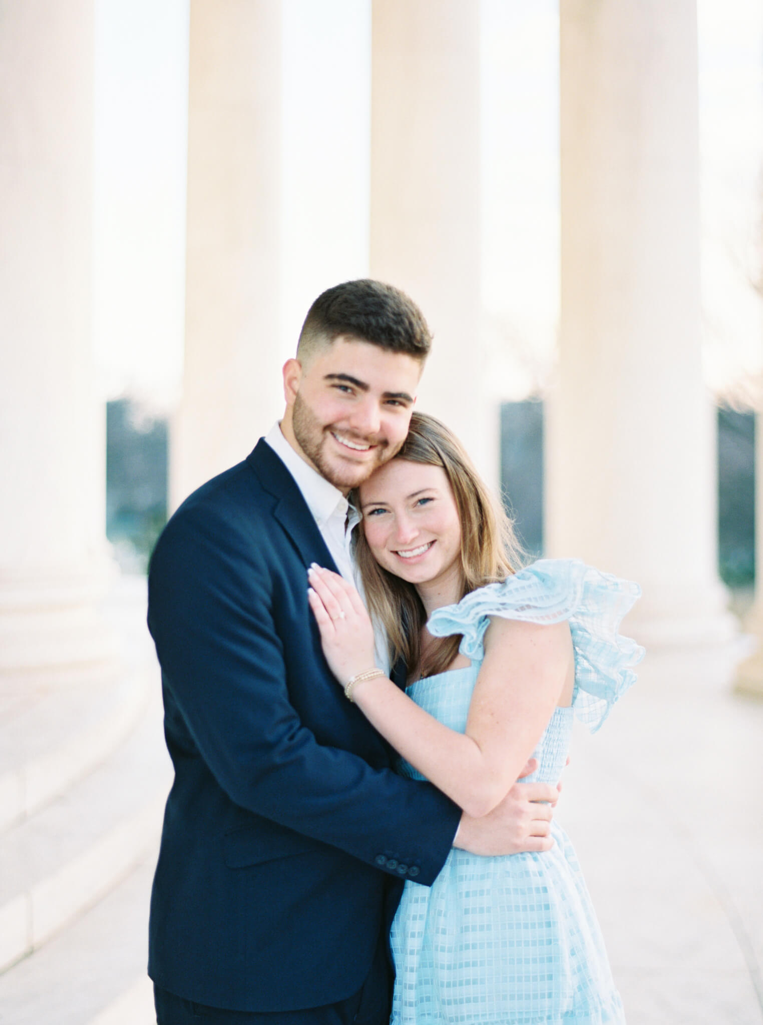 A young man in a navy blue suit holding his fiancée in a light blue dress close during their Jefferson Memorial Engagement Photos with marble columns in the background