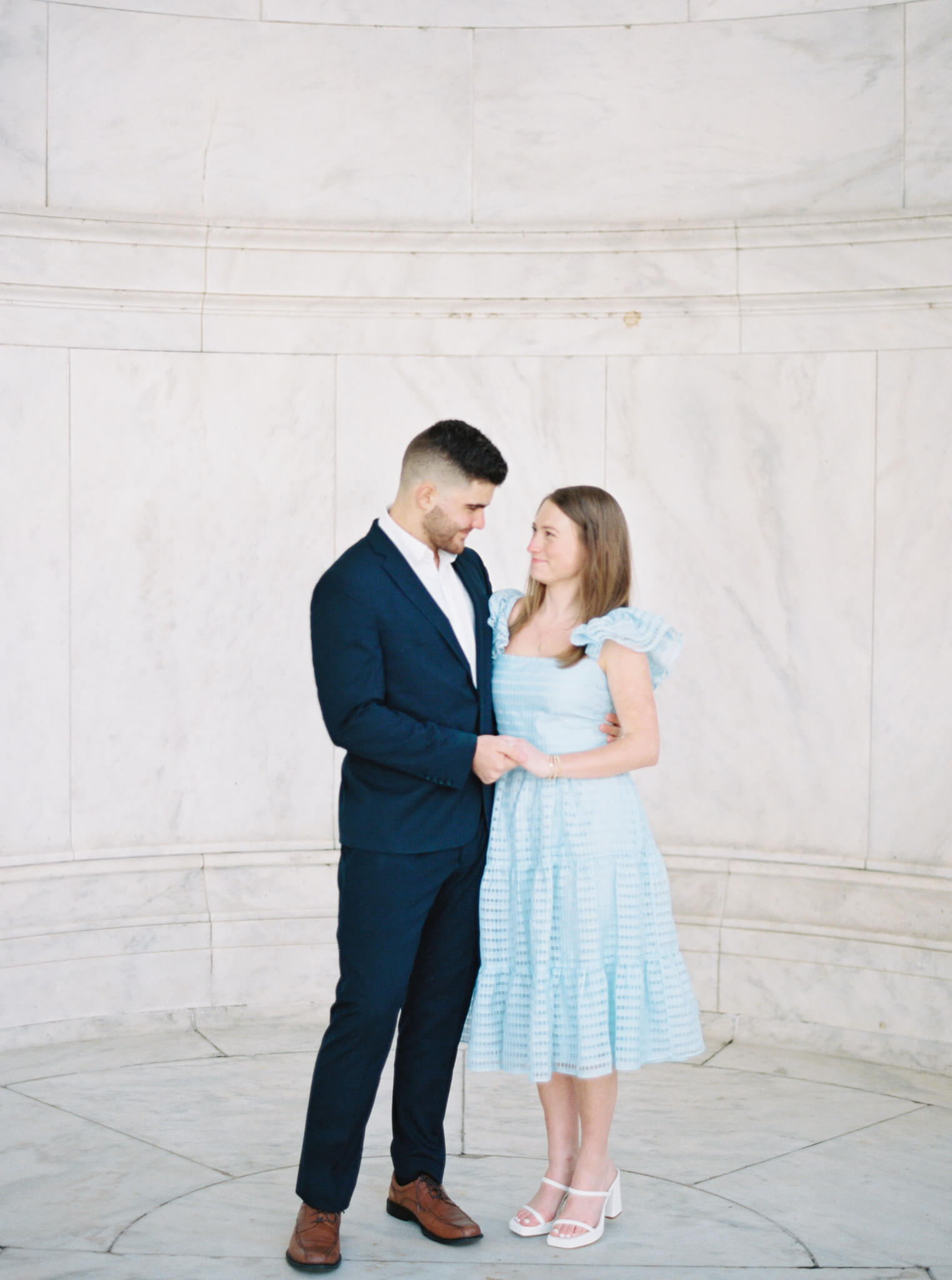 A man in a blue suit and his fiancée wearing a light blue dress standing arm in arm in front of a marble wall at the Jefferson Memorial looking at each other