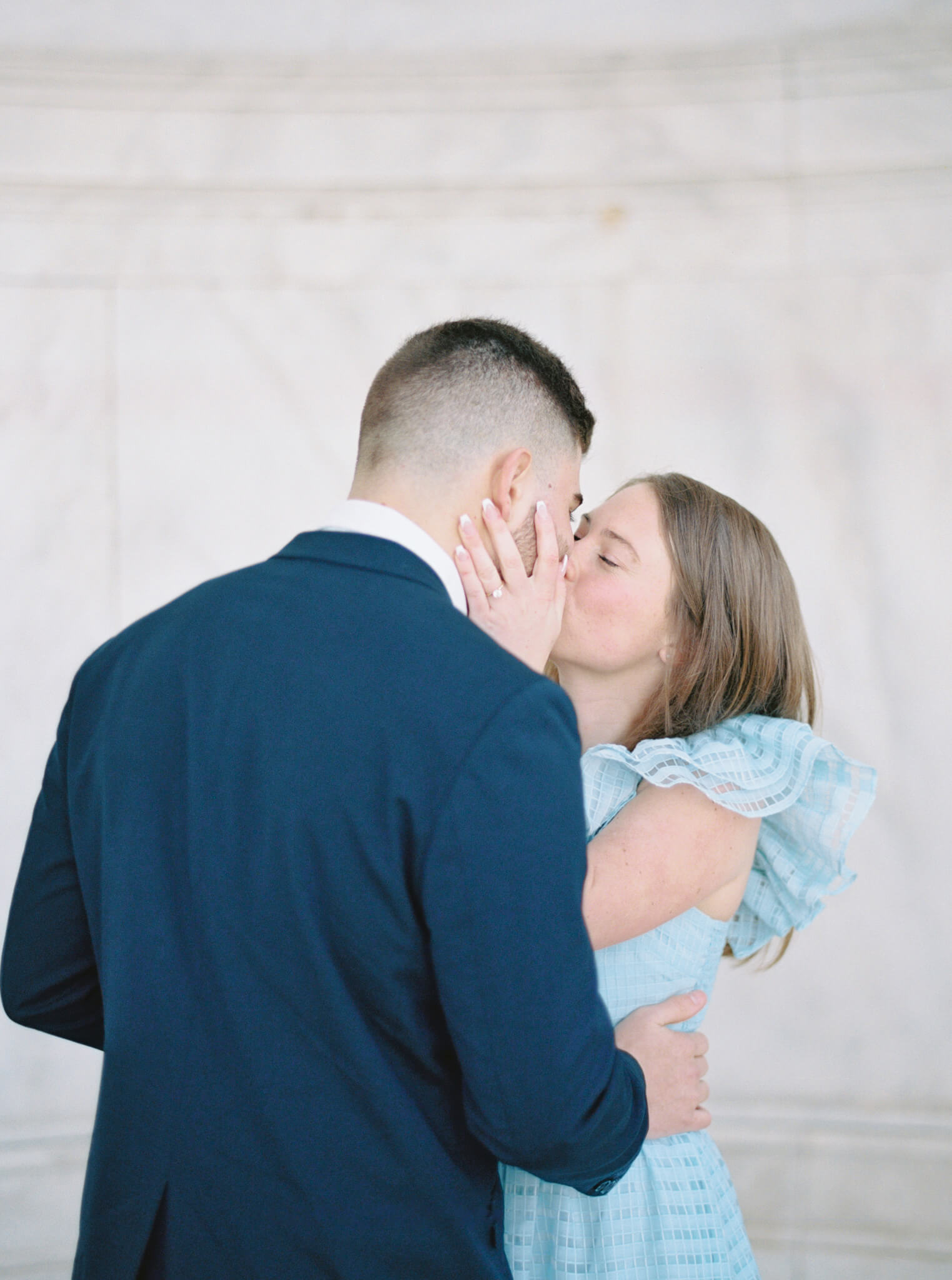Closeup of a man in a navy suit with his back turned, being kissed by a young woman in a light blue dress during their engagement session