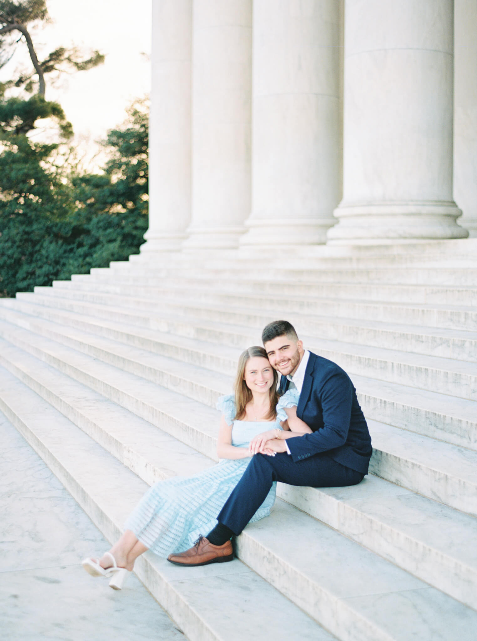 An engaged couple wearing a navy blue suit and light blue dress sitting on the steps of the Jefferson Memorial during their engagement session