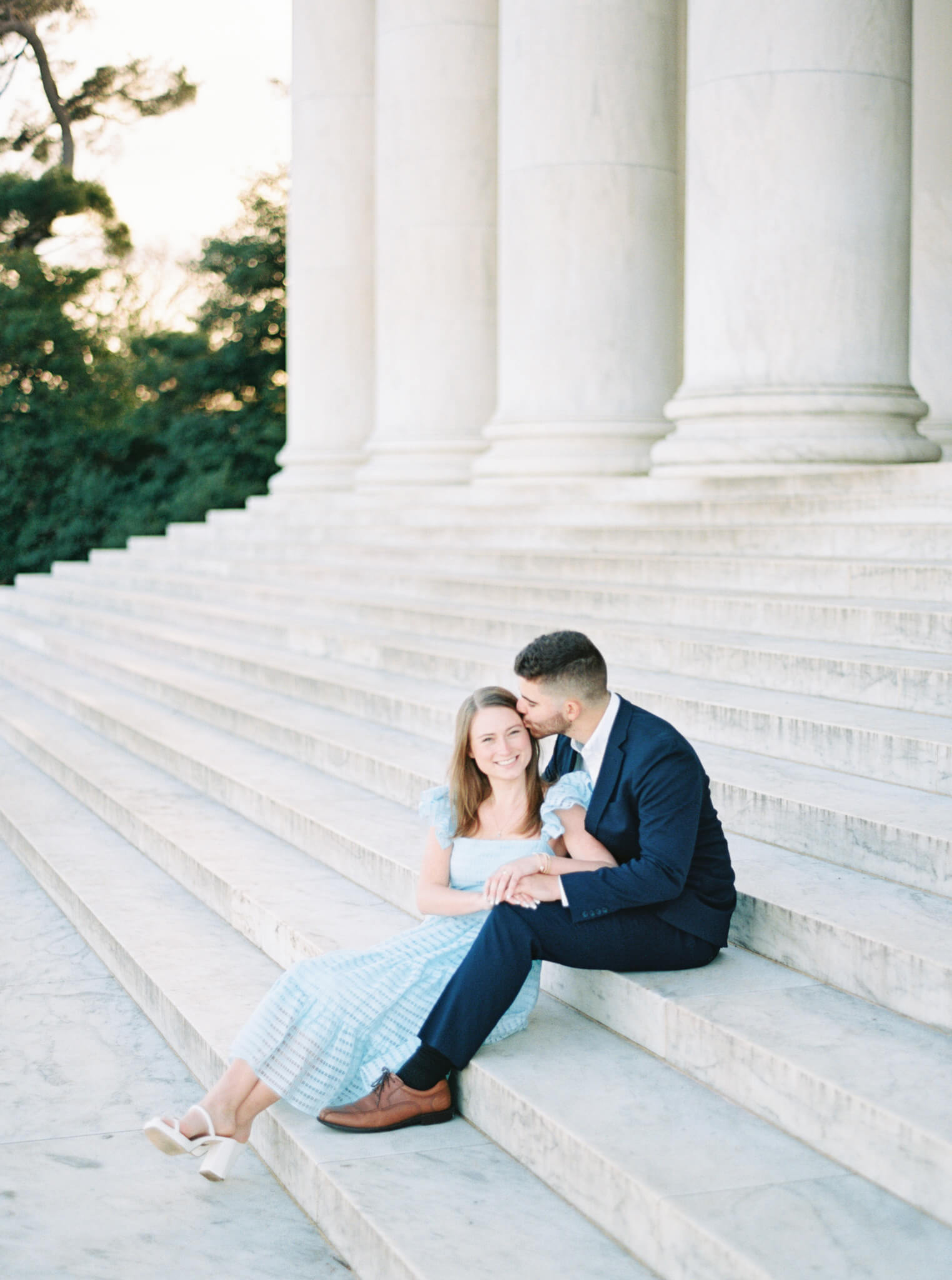 A man in a navy suit and a woman in a light blue dress sitting on the steps of the Jefferson Memorial while he kisses her temple 