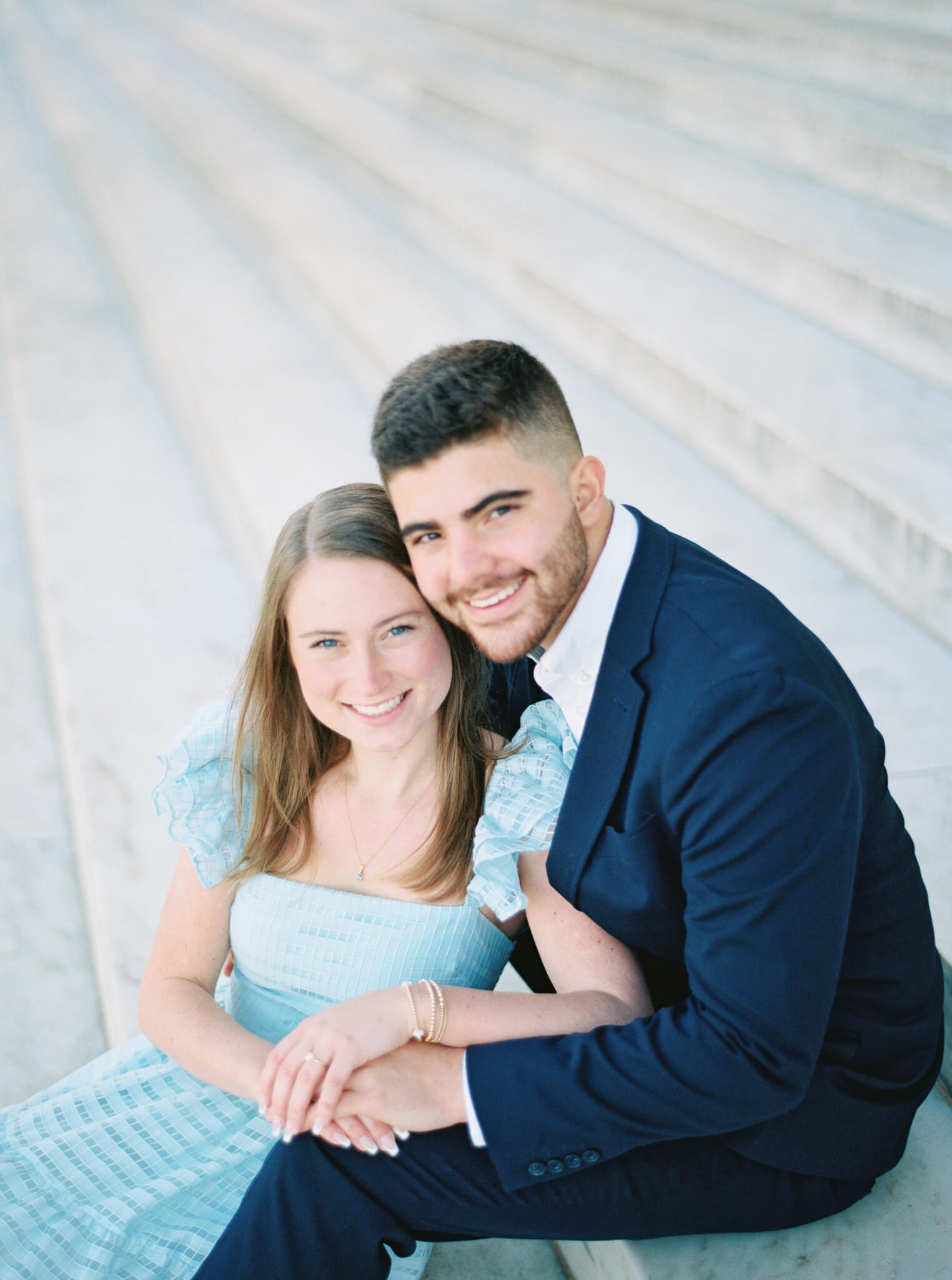 Closeup of an engaged couple in a navy blue suit and light blue tulle dress sitting on the steps of the Jefferson Memorial holding each other close