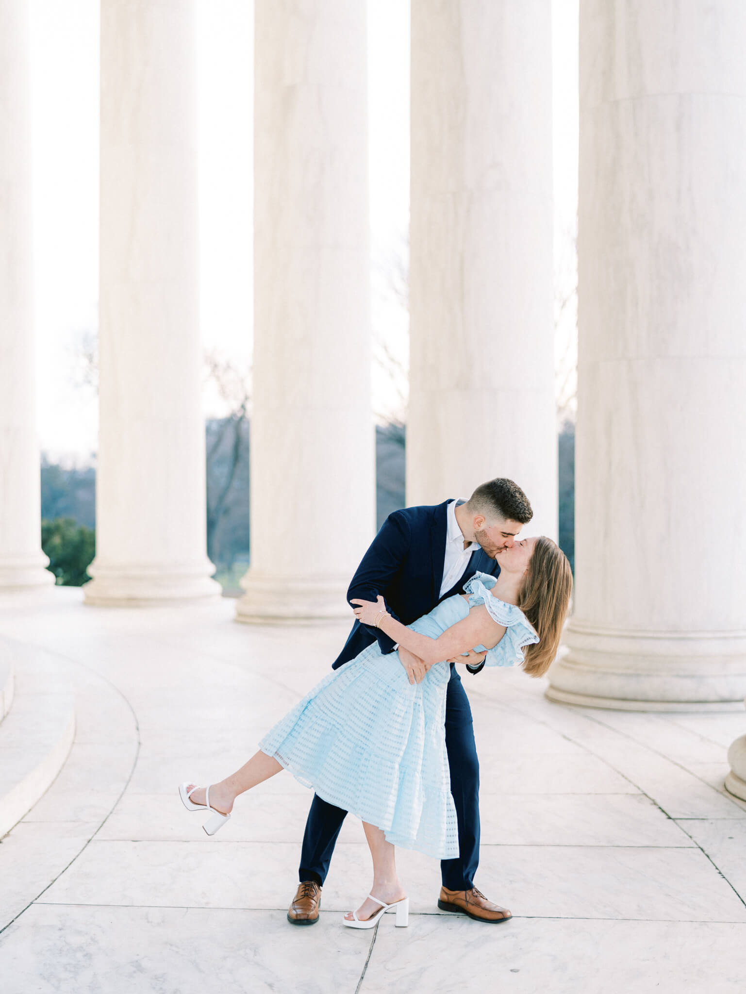 A man in a navy blue suit dipping back and kissing his fiancée wearing a light blue dress in front of the marble columns of the Jefferson Memorial.