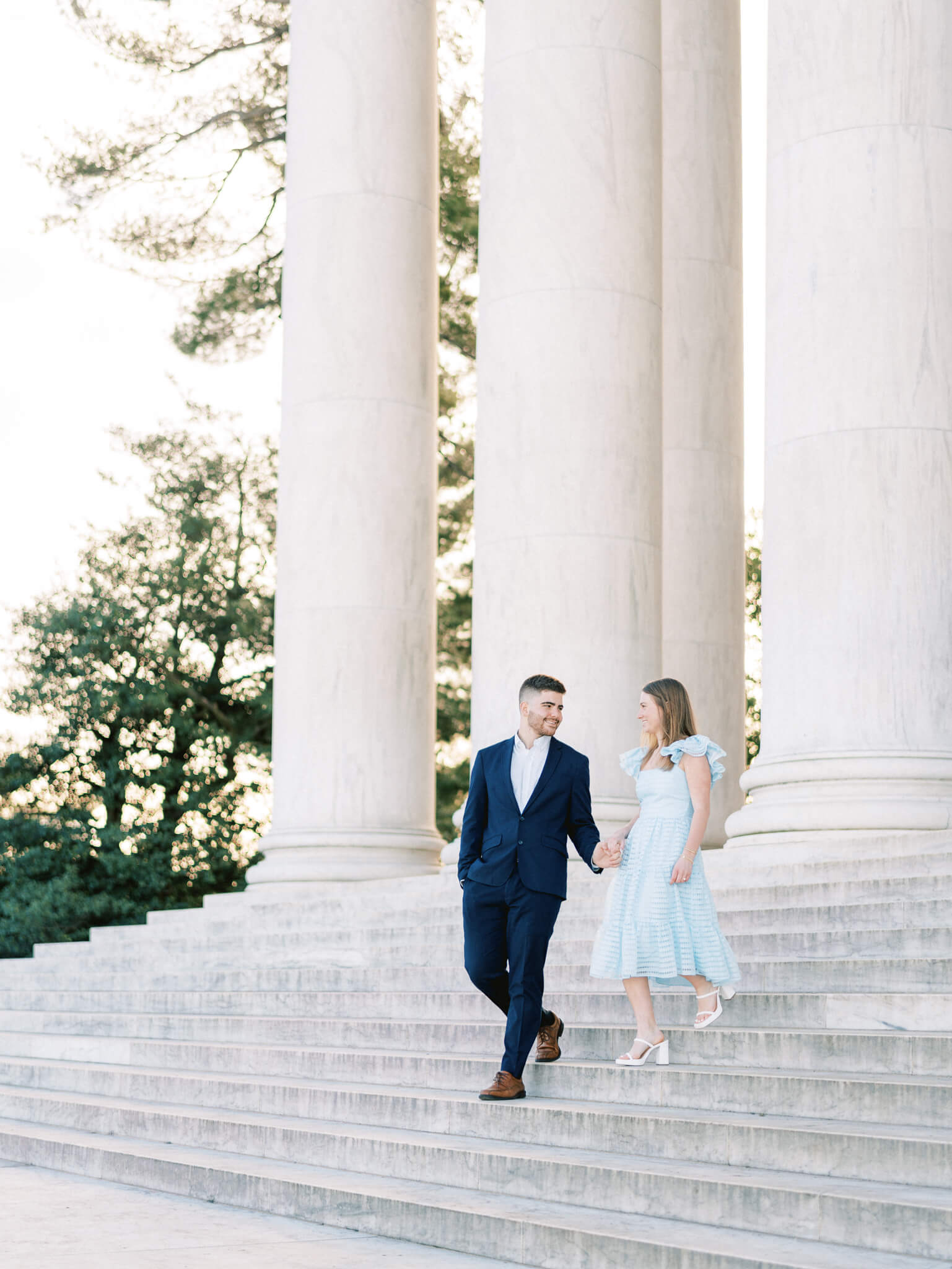 A man in a navy blue suit and a woman in a light blue dress walking down the steps of the Jefferson Memorial while looking at each other with columns in the background.