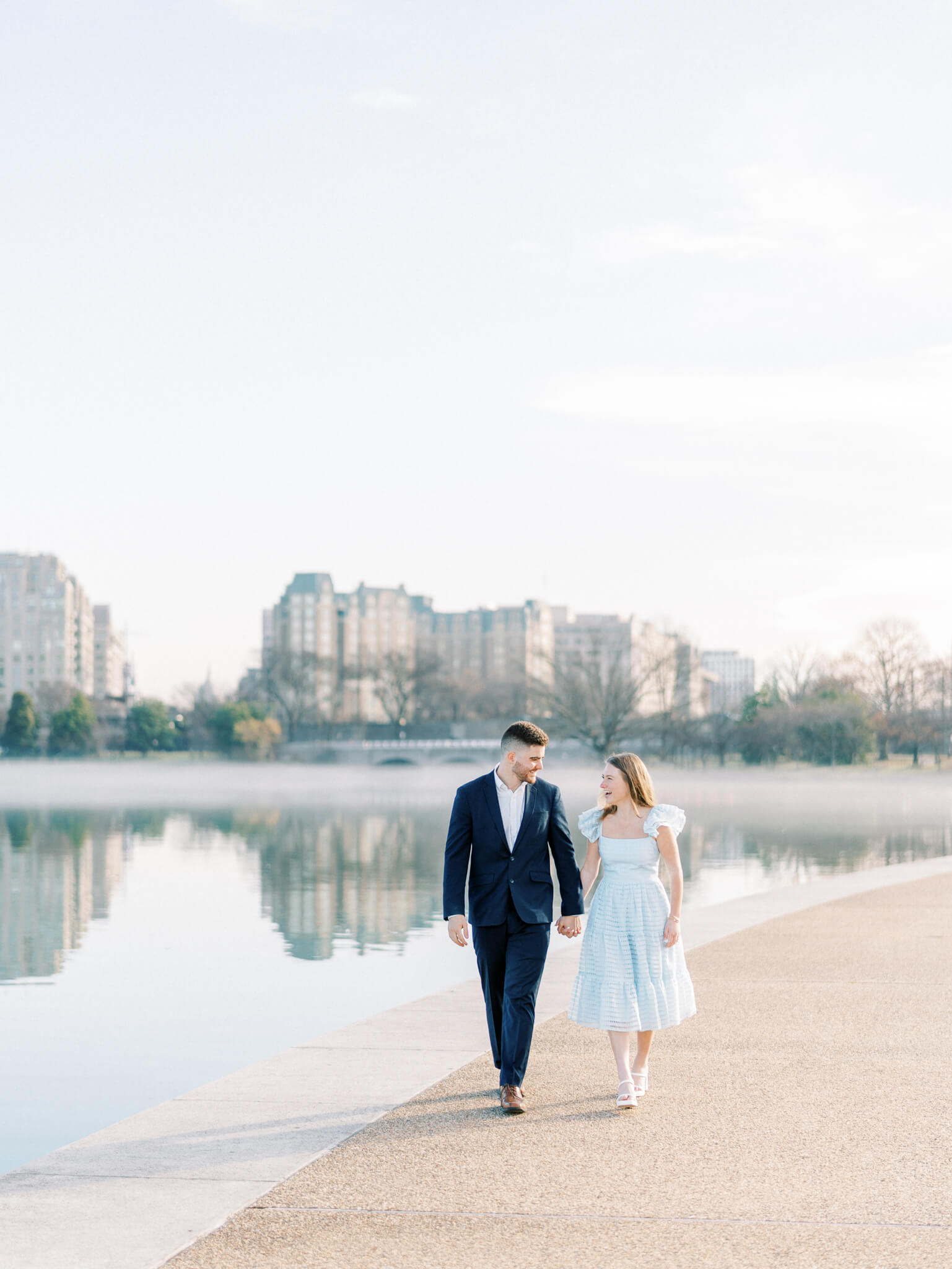 A man in a navy blue suit and a woman in a light blue dress walking on the edge of the Tidal Basin for their Jefferson Memorial Engagement Photos.