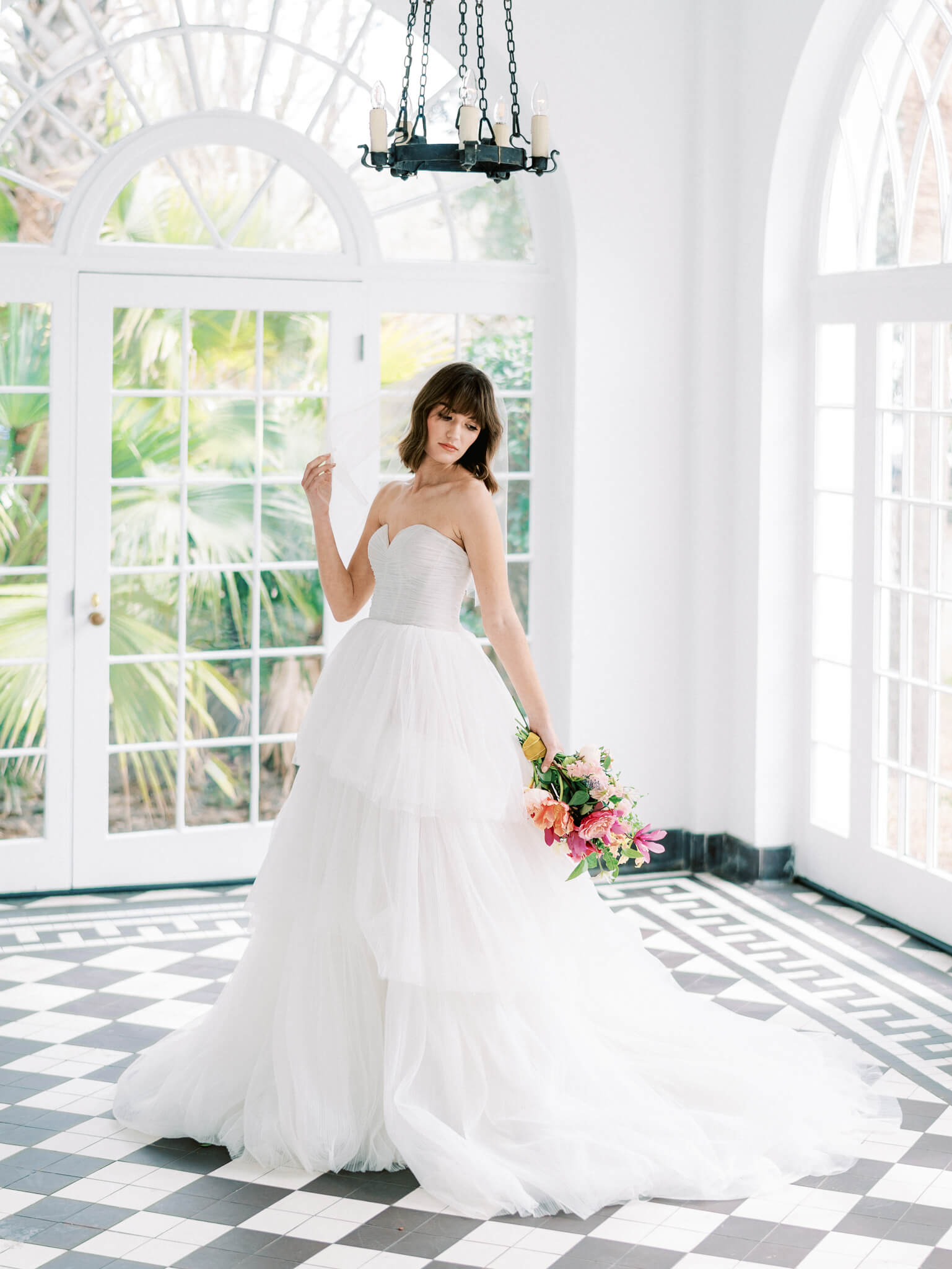 Portrait of a bride in a tulle gown holding her bouquet inside Lowndes Grove's entry with arched glass windows and black and white checkered floors.