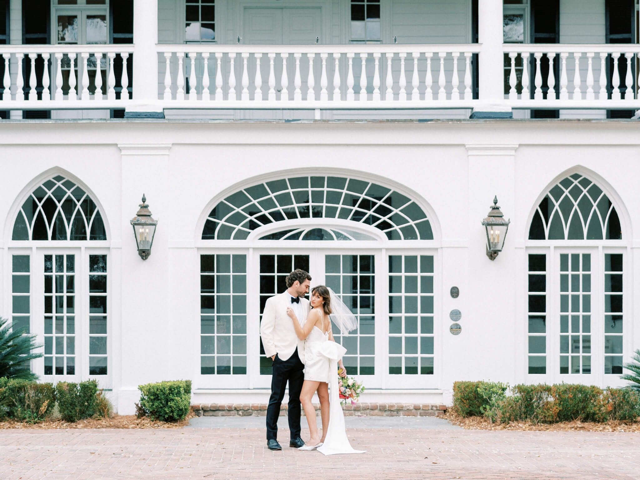 A bride and groom embracing in front of Lowndes Grove's arched windows and porch.
