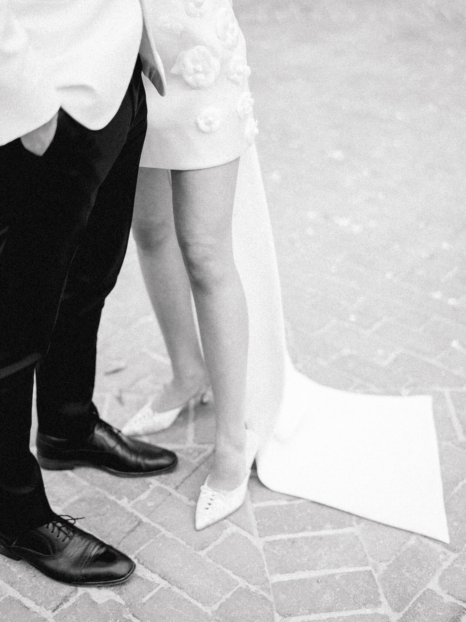 A bride and groom's legs and feet standing closely together wearing a black and white tux and short wedding dress.