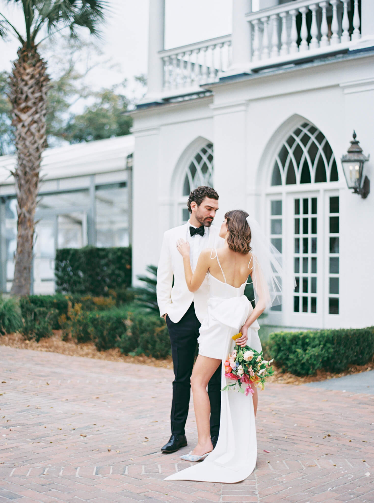 A bride in a short dress and groom in a tux embracing in front of Lowndes Grove wedding venue.