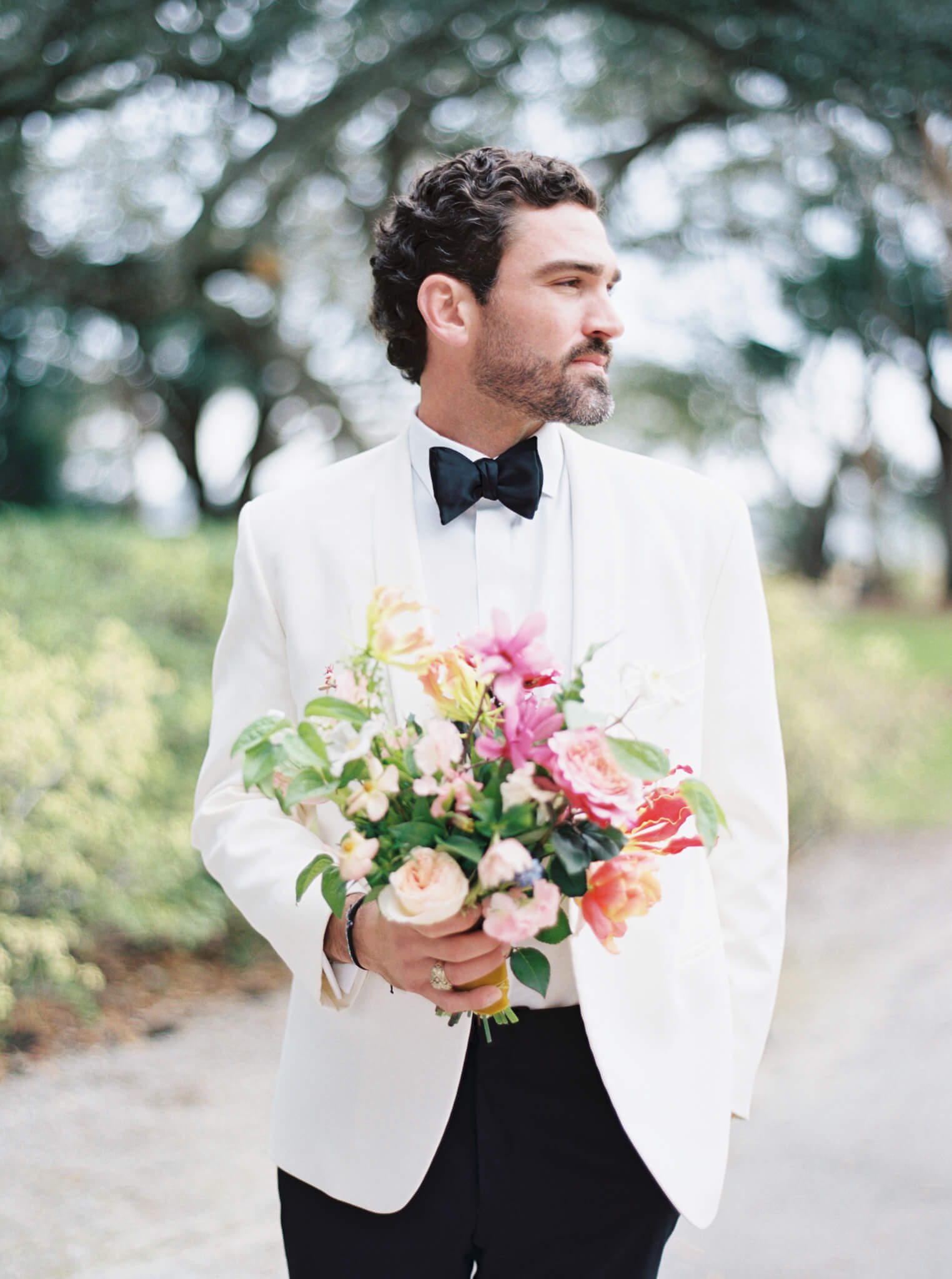 Closeup of a groom in a white and black tuxedo looking to the side while holding a pink and yellow bouquet of flowers.