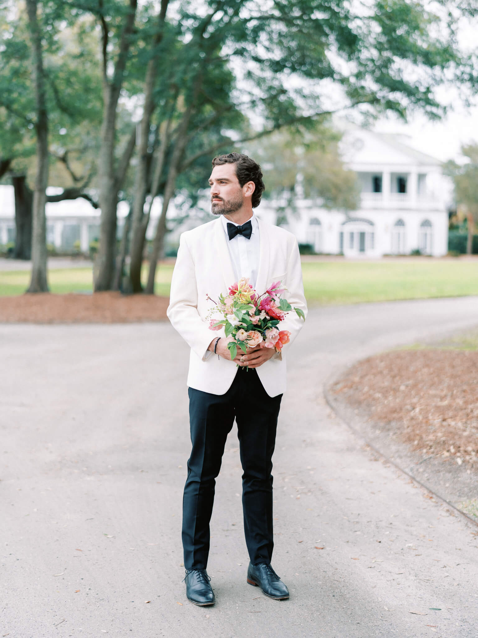 A groom holding a bouquet and wearing a d black and white tux standing in front of Lowndes Grove Wedding Venue.