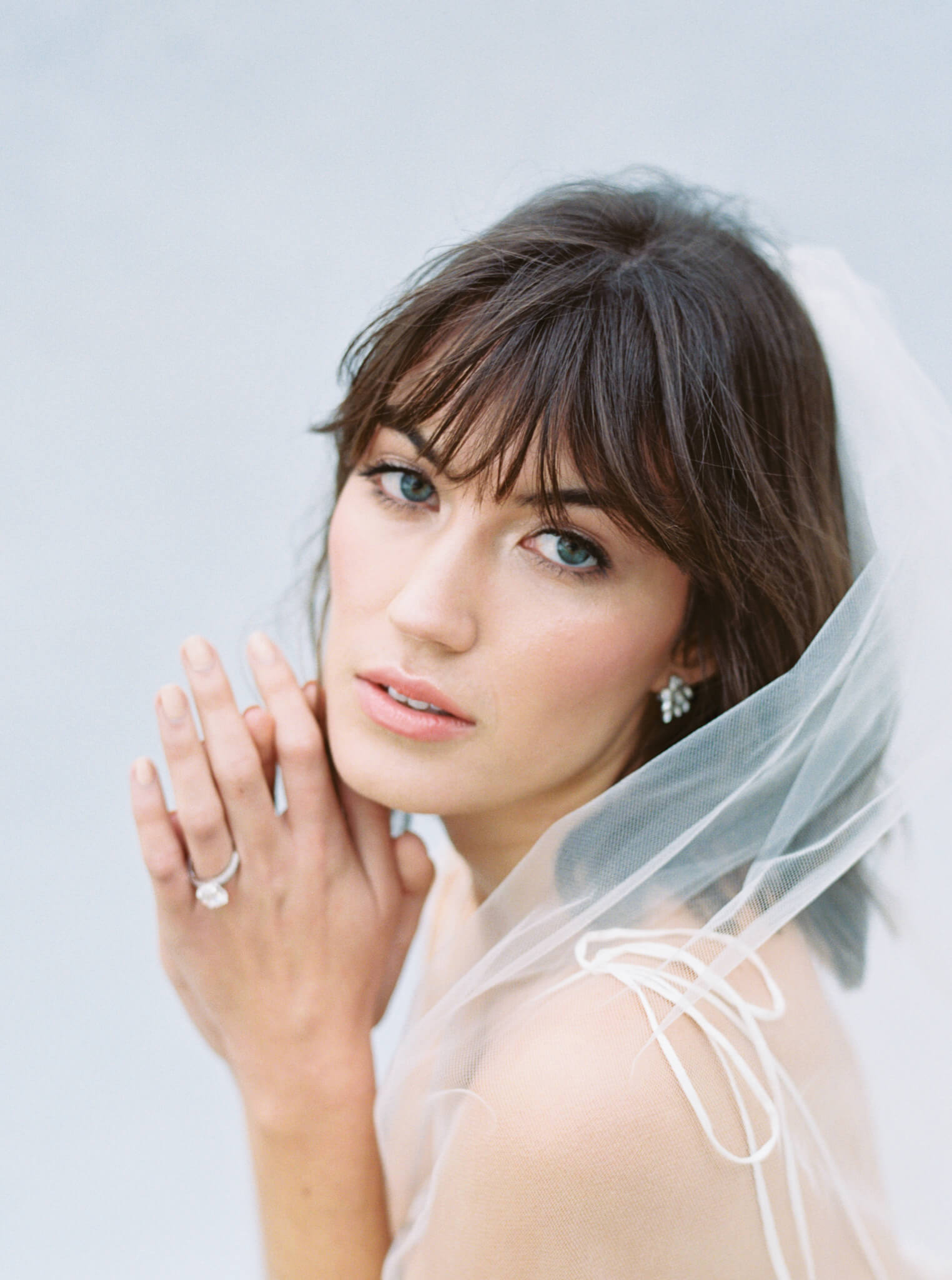 Closeup of a brown haired bride wearing a veil and holding her hands to the side of her face in front of a blue backdrop.