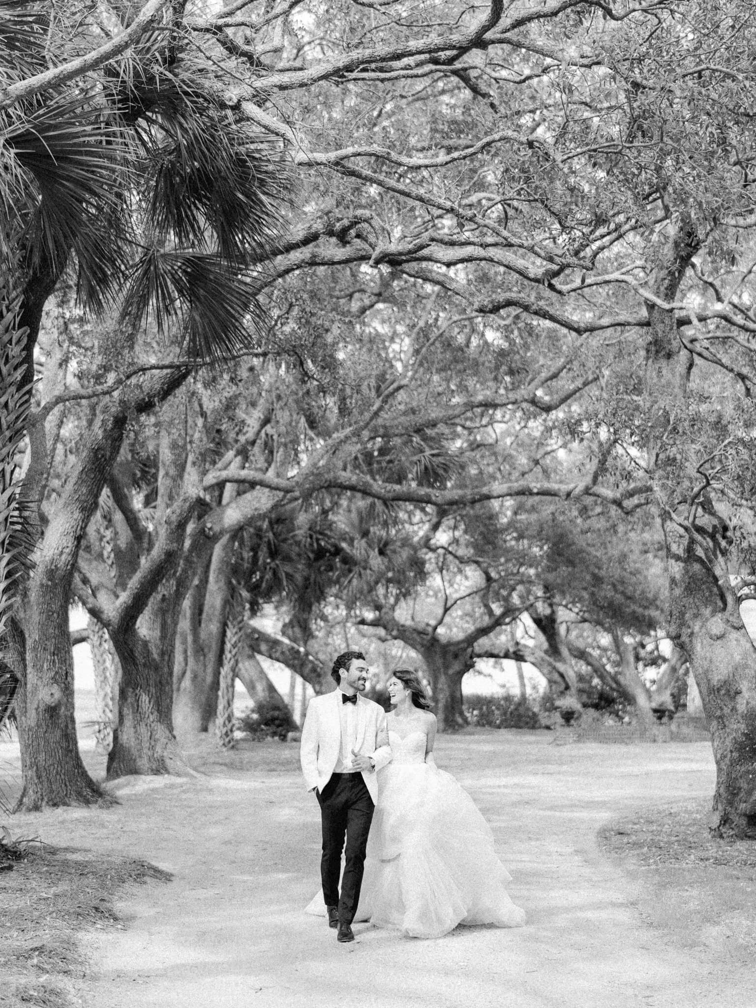 Black and white image of a bride and groom walking with linked arms and looking at each other under a canopy of large live oak trees at Lowndes Grove Wedding Venue.