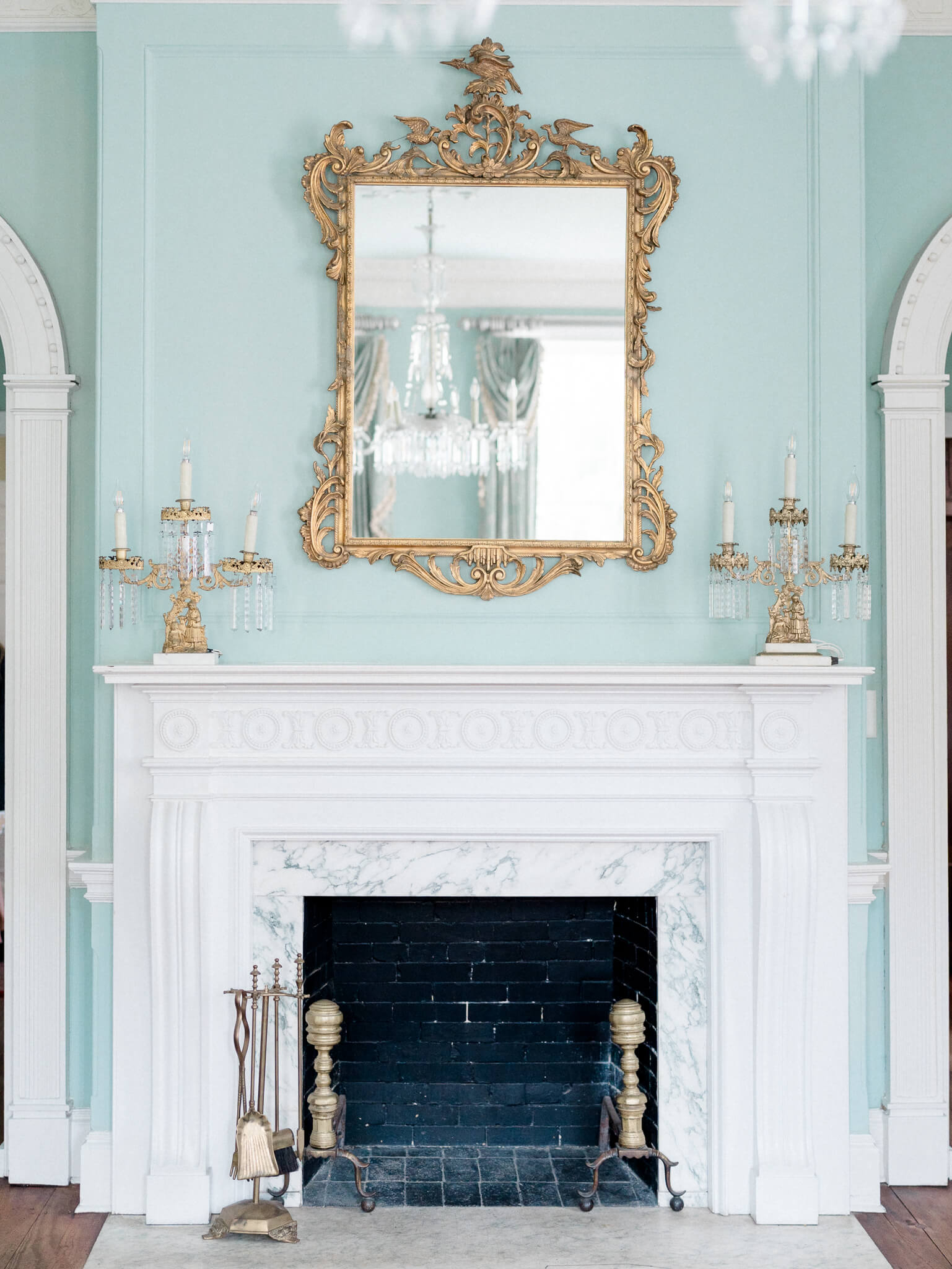A white fireplace with a large gold mirror above and turquoise walls at Lowndes Grove Wedding Venue.