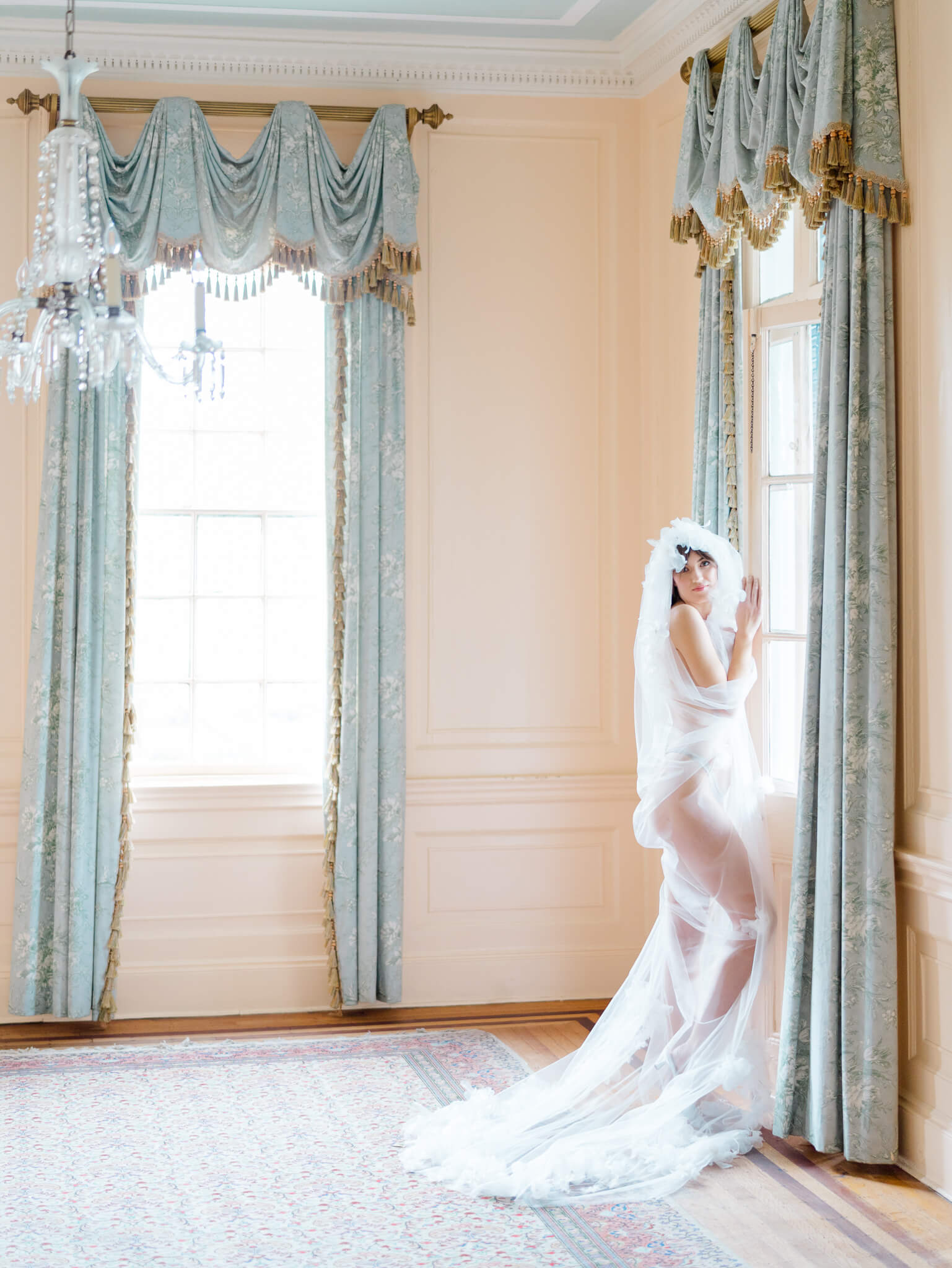 A bridal boudoir image of a bride covered in only her veil standing by the window in Lowndes Grove's pink room.