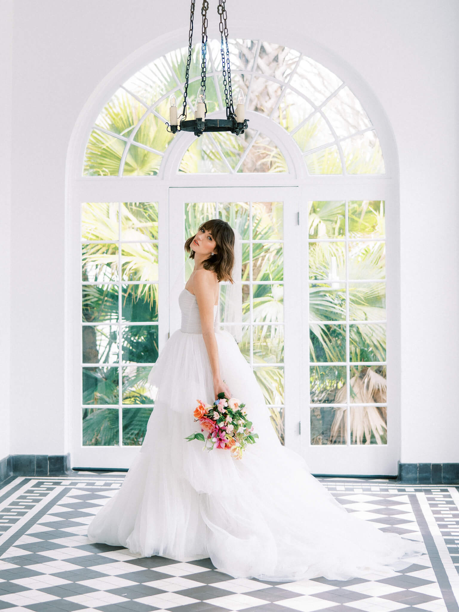 A bride in a tulle gown holding a bouquet of bright flowers standing on a checkered black and white floor in front of an arched window.
