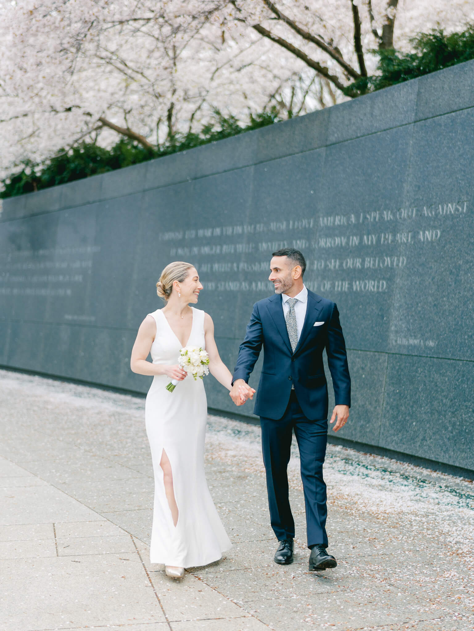 A bride and groom walking hand in hand while looking at each other under the cherry blossom trees at the MLK Memorial in Washington, D.C.