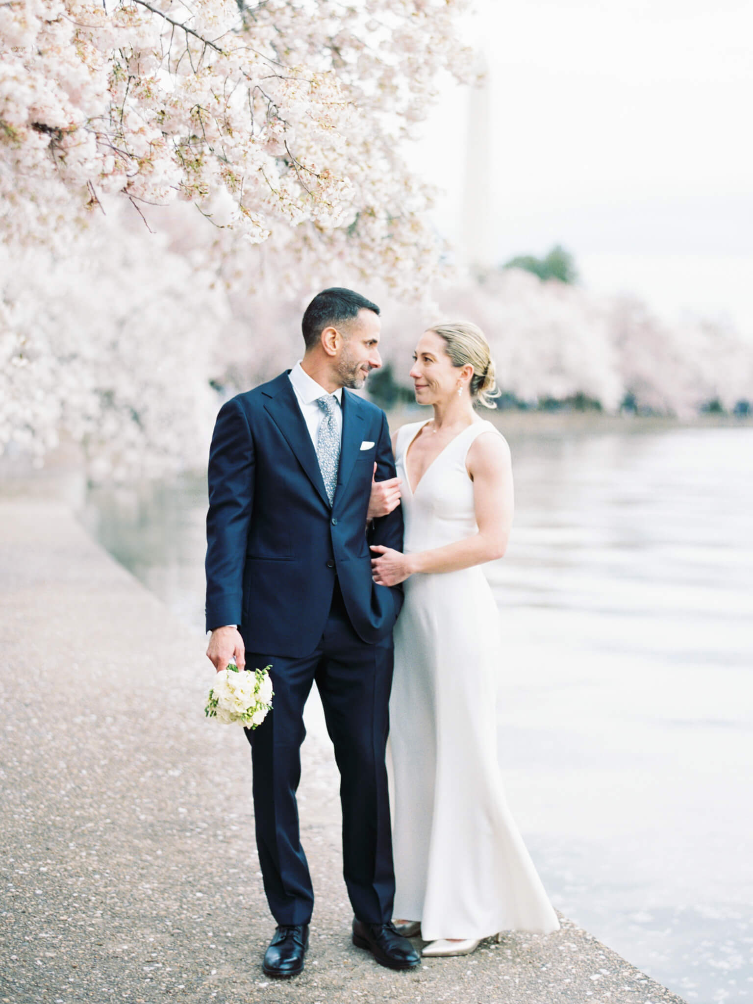 A bride and groom looking at each other while he holds her bouquet in front of the cherry tree lined tidal basin