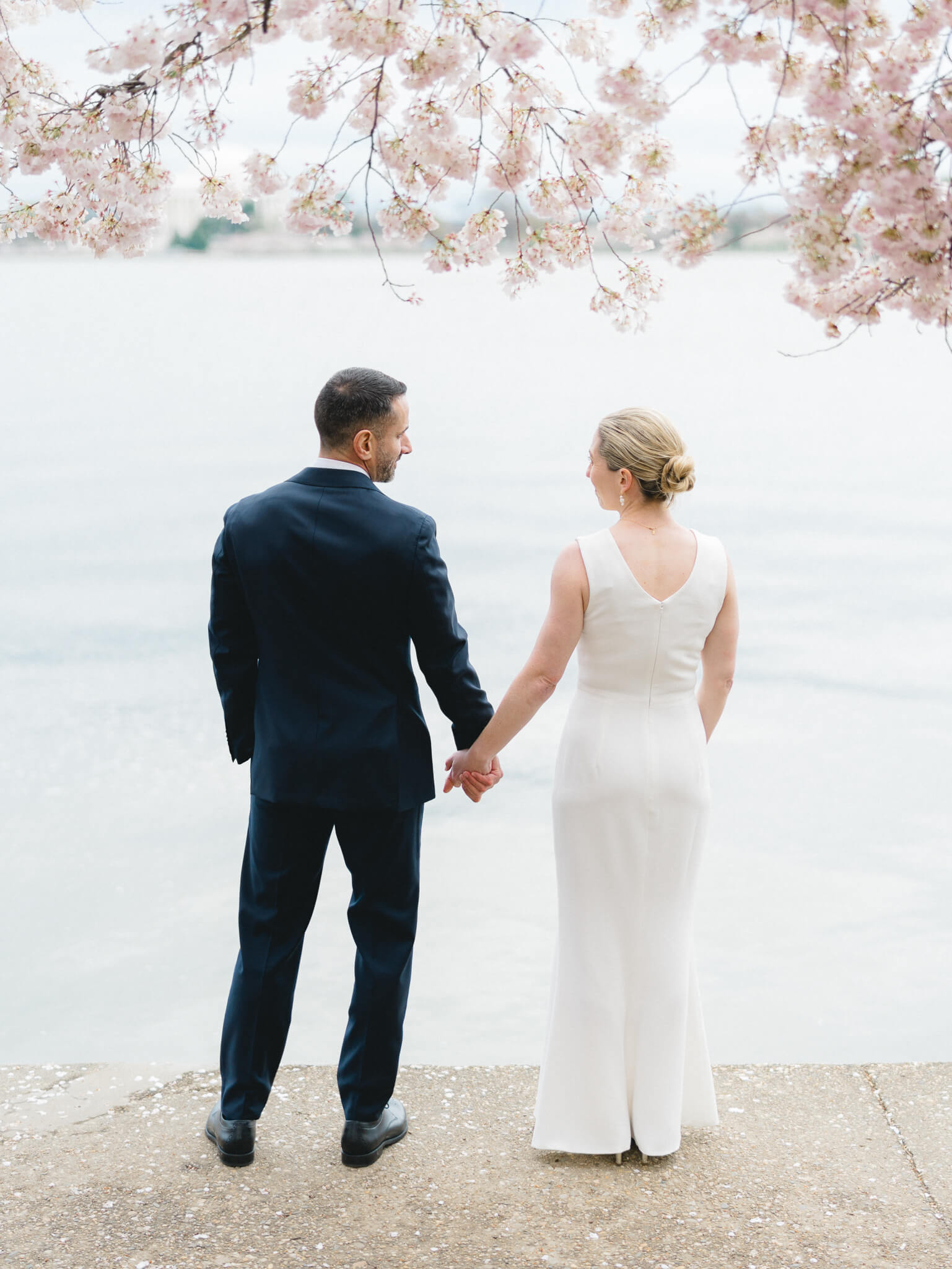A bride and groom with their backs turned looking out over the tidal basin with cherry blossoms above them