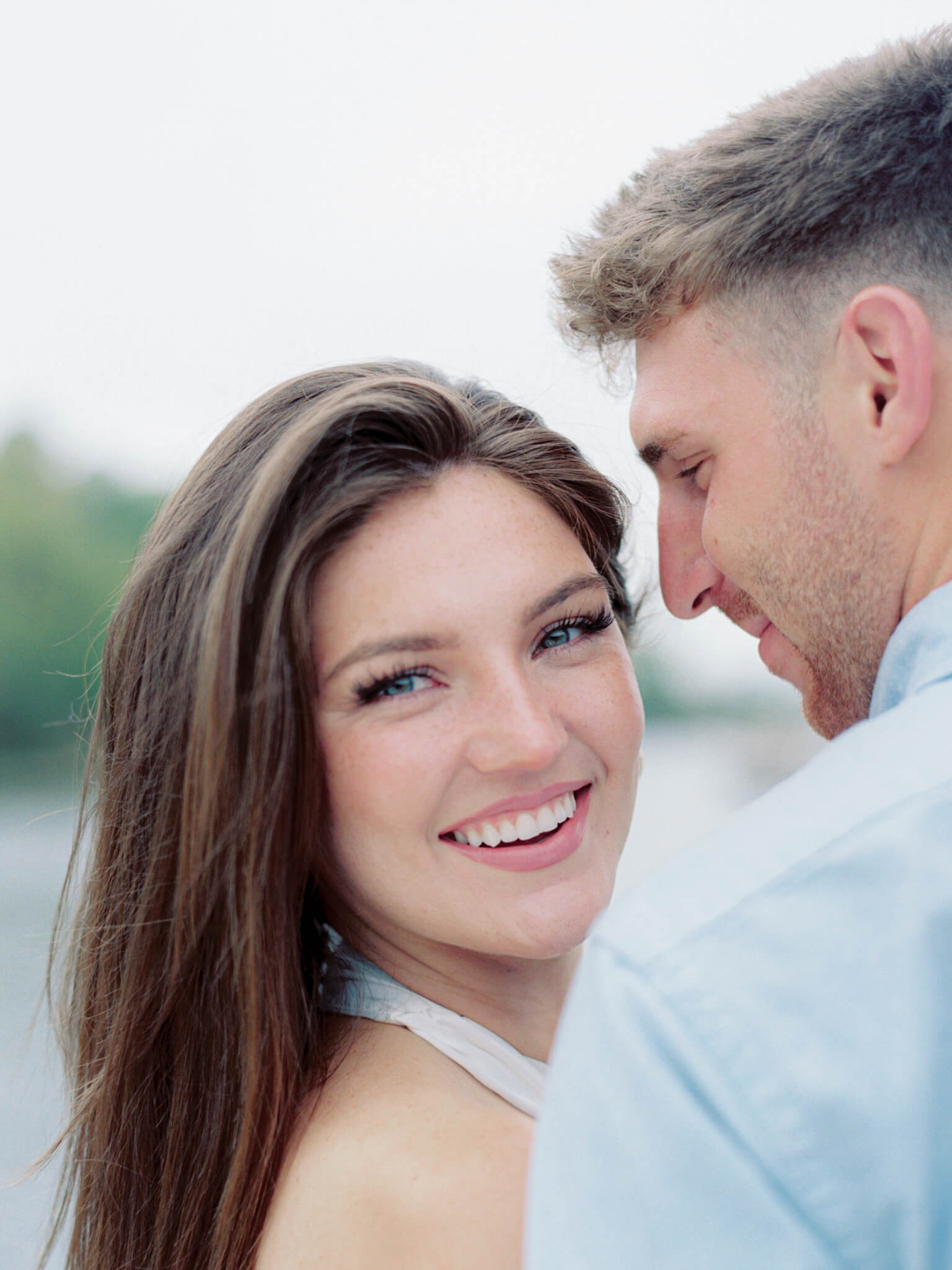 Closeup of a woman looking at the camera while her fiancé looks at her with water in the background.
