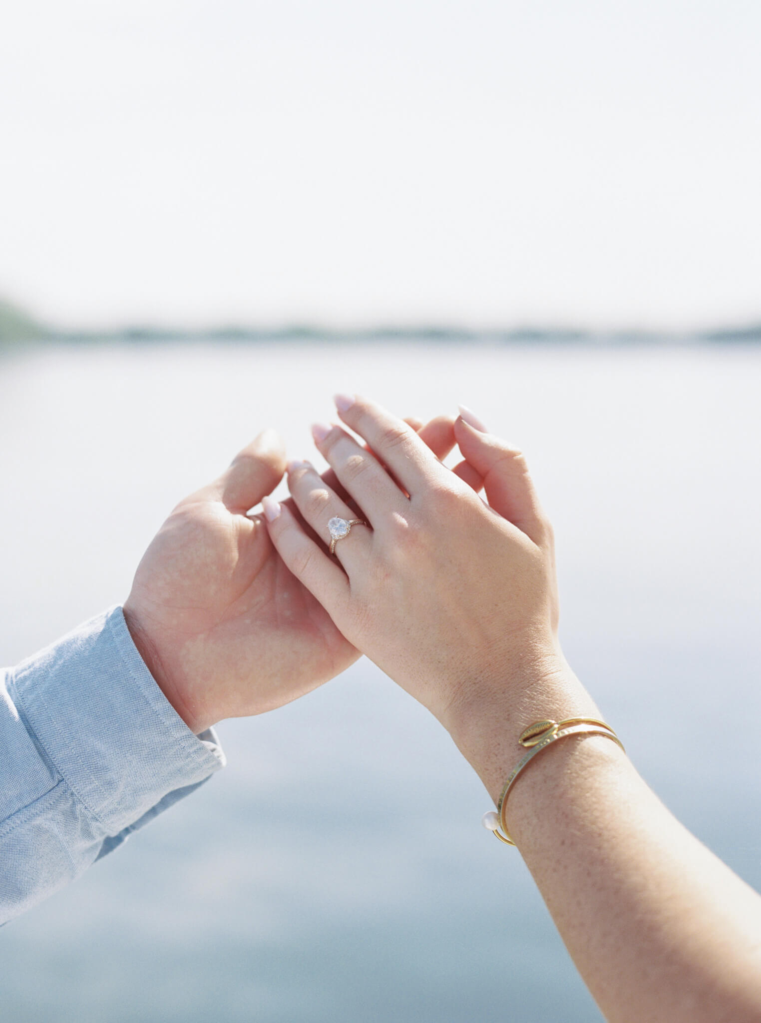 Closeup image of a man's hand holding a woman's hand showing off an engagement ring with water in the background.