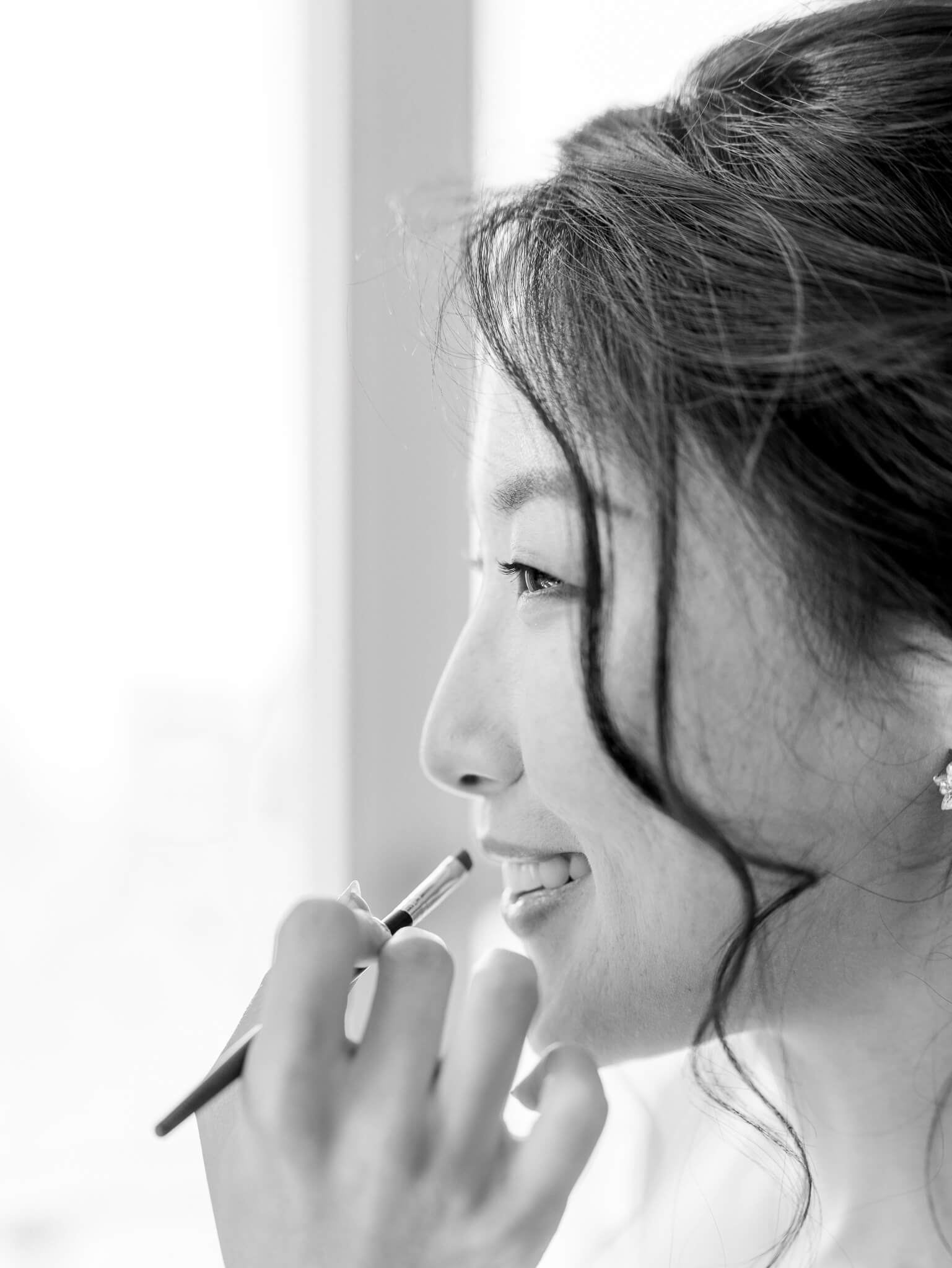 Black and white closeup image of a bride having her lipstick applied with a brush.