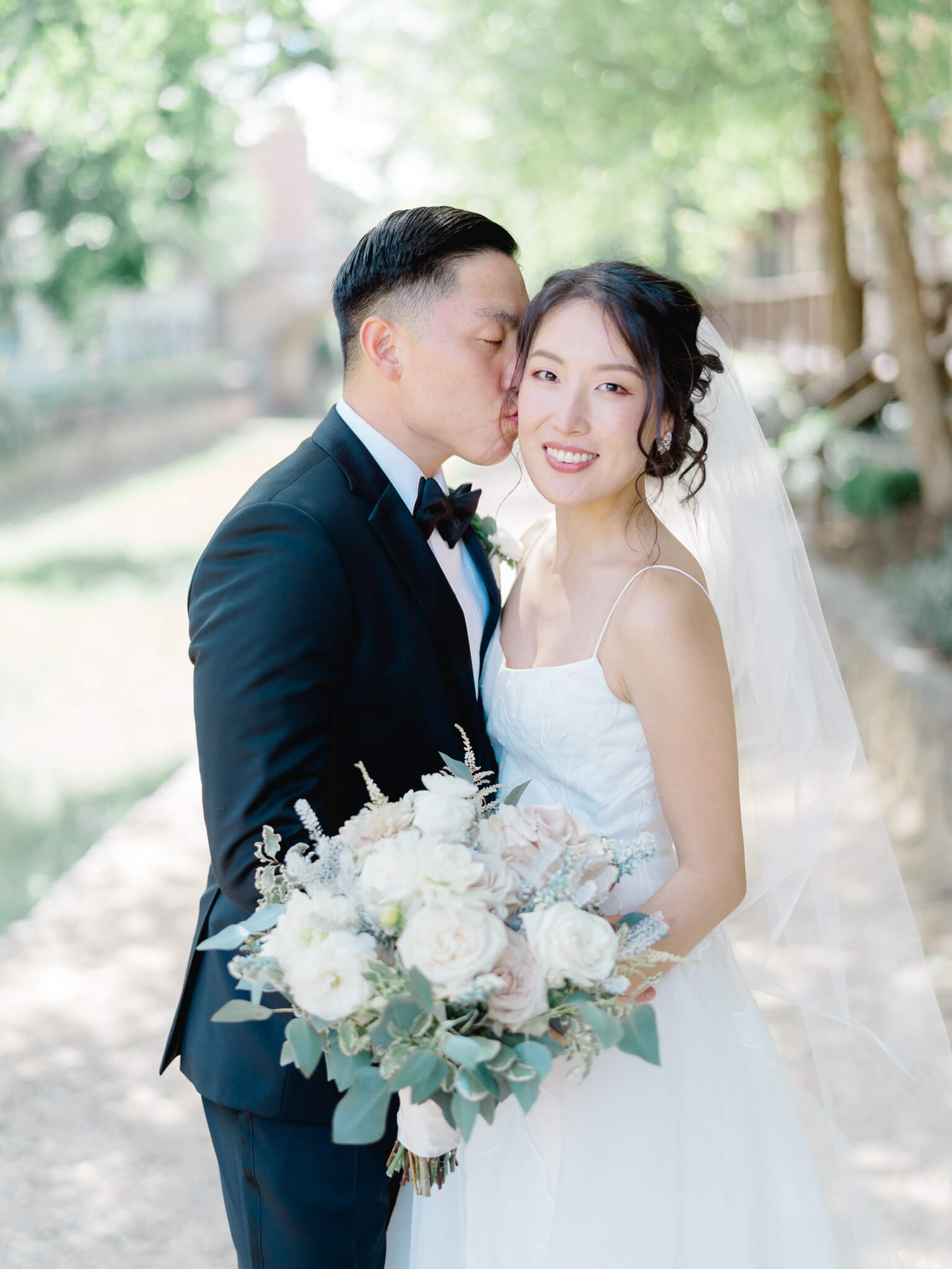 A groom kissing his bride on the cheek while she looks at the camera and holds her bouquet at their Westin Georgetown Wedding.