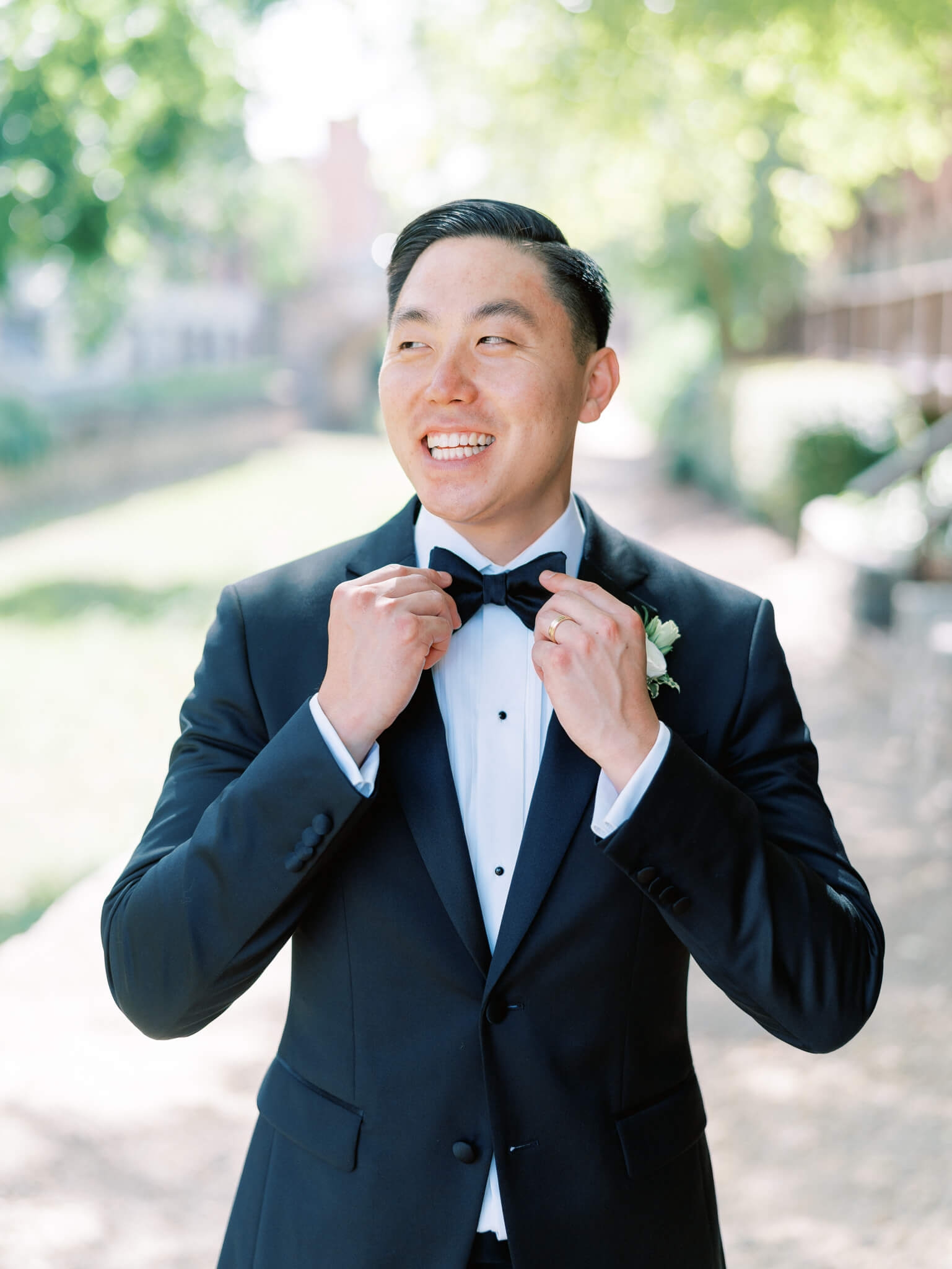 A groom straightening his bow tie with his hands while smiling off to the side on his wedding day.