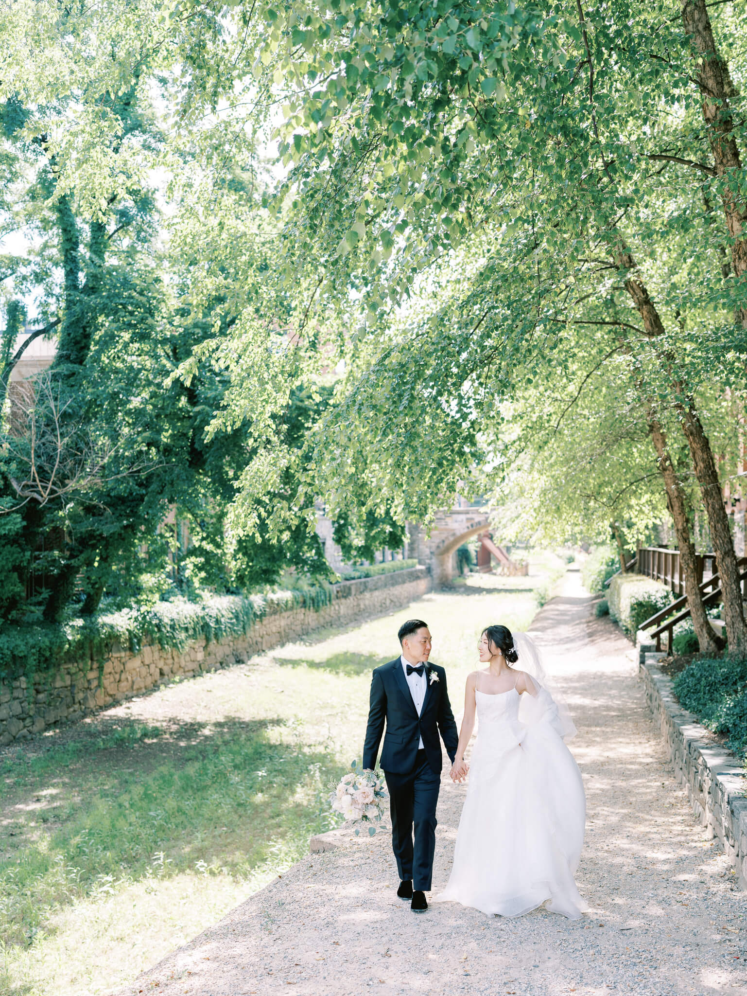 A bride and groom walking hand in hand next to a dry canal under green trees in Georgetown, D.C.