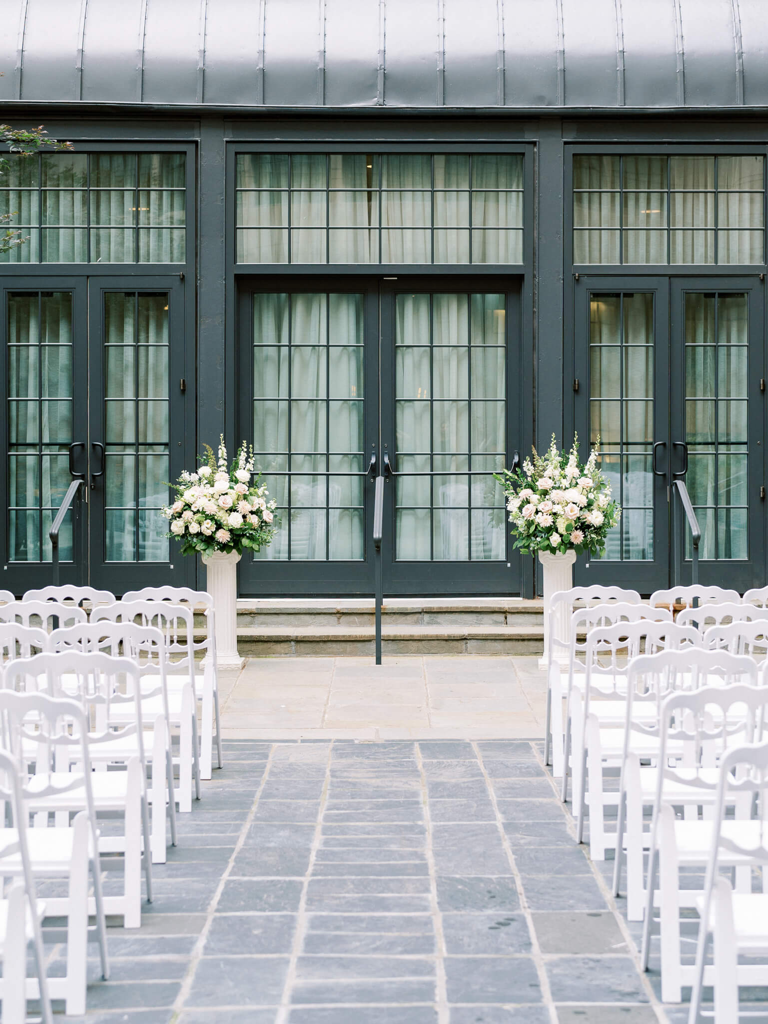 A large black glass window inside the courtyard at the Westin Georgetown with white ceremony chairs and floral centerpieces in front of it.