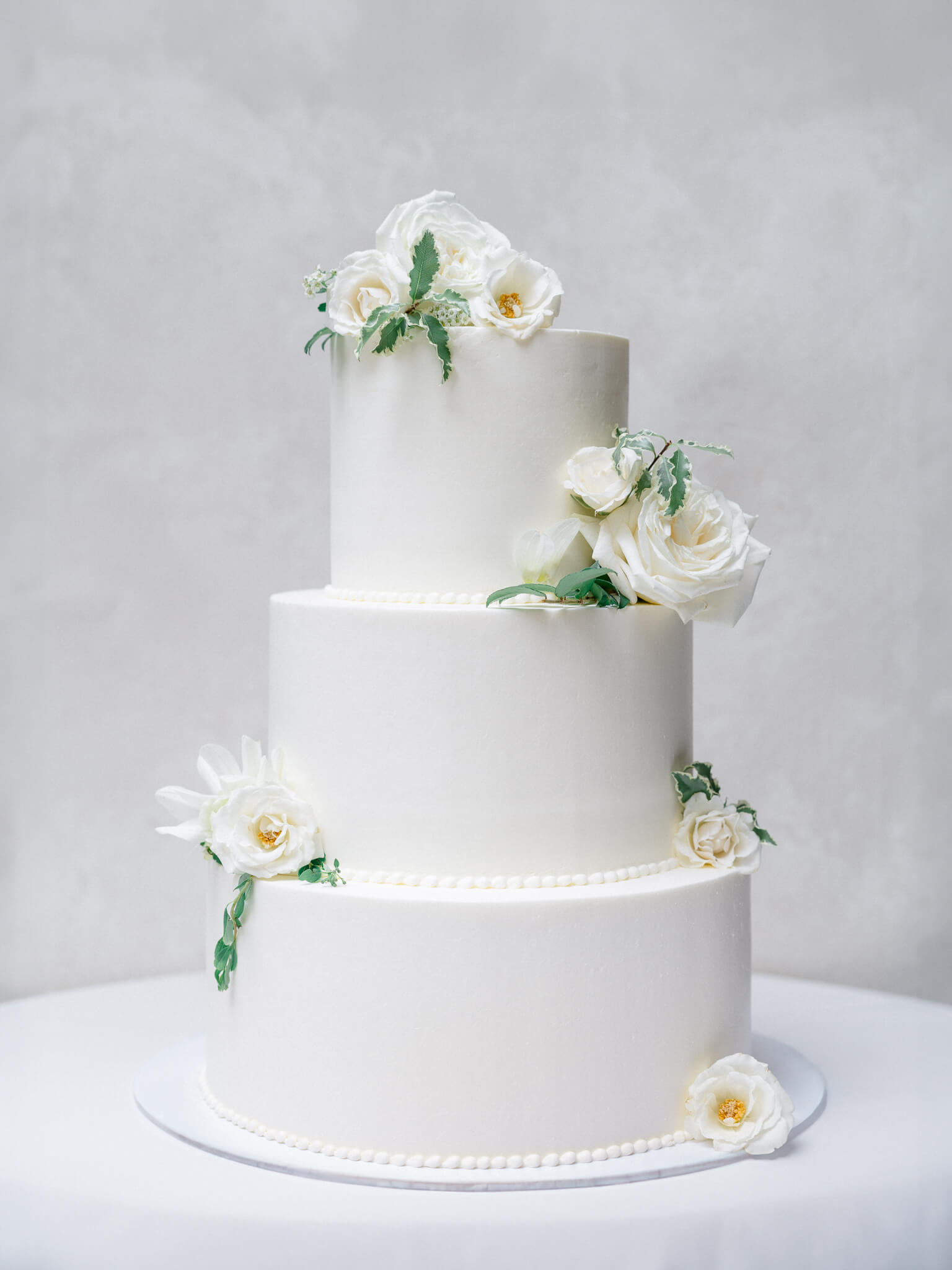 A white three tier wedding cake with greenery and cream roses sitting in front of a grey backdrop.