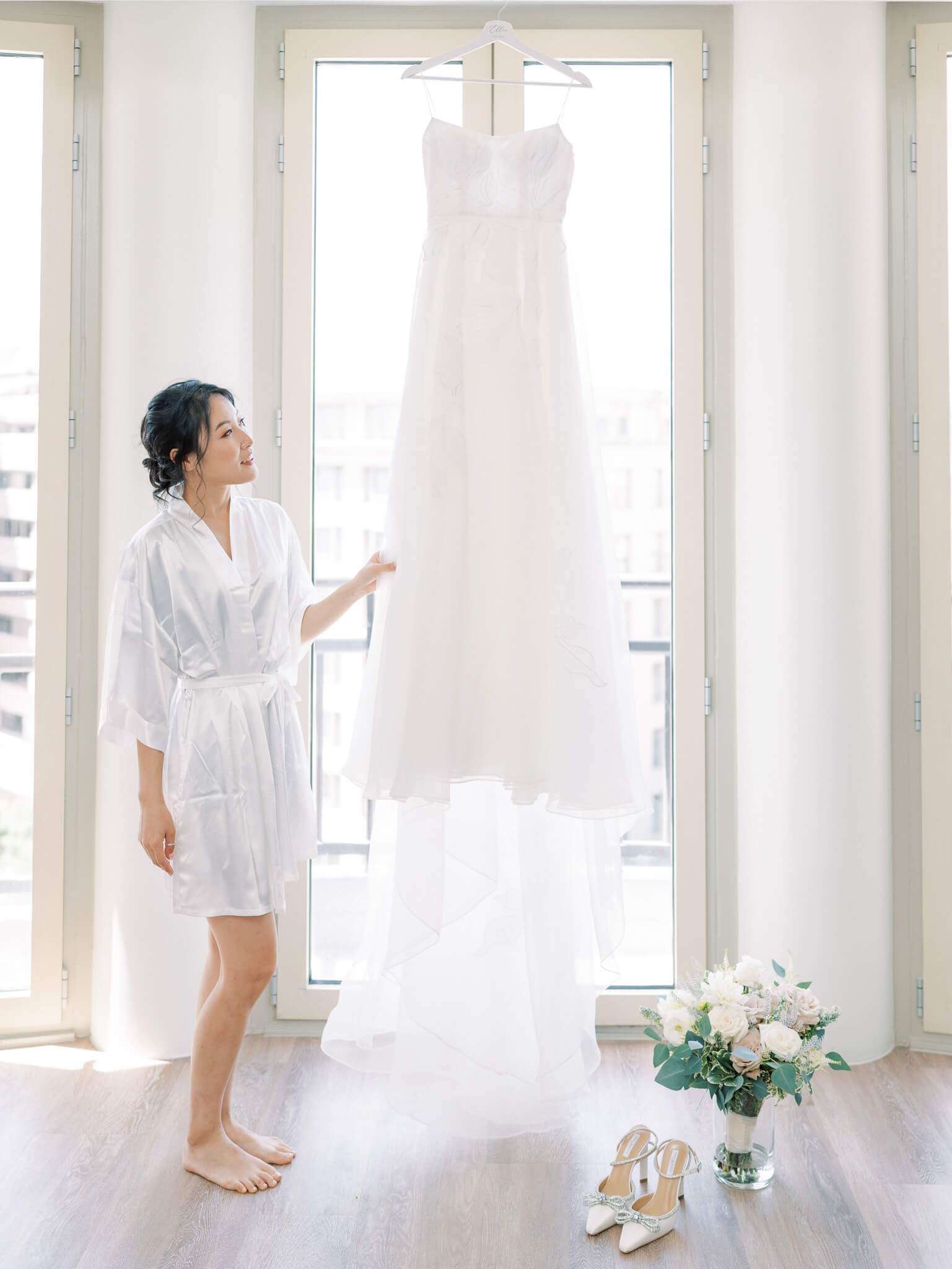 A bride in a white robe touching her wedding gown, which is hanging in front of a window.