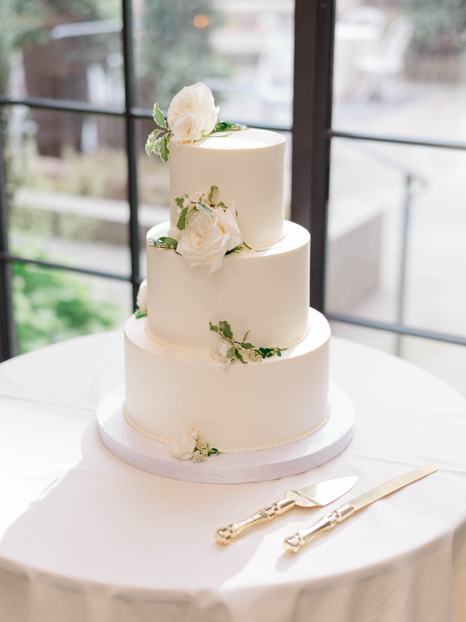 A white three tier wedding cake with cream roses and greenery sitting on a table at a Westin Georgetown Wedding.