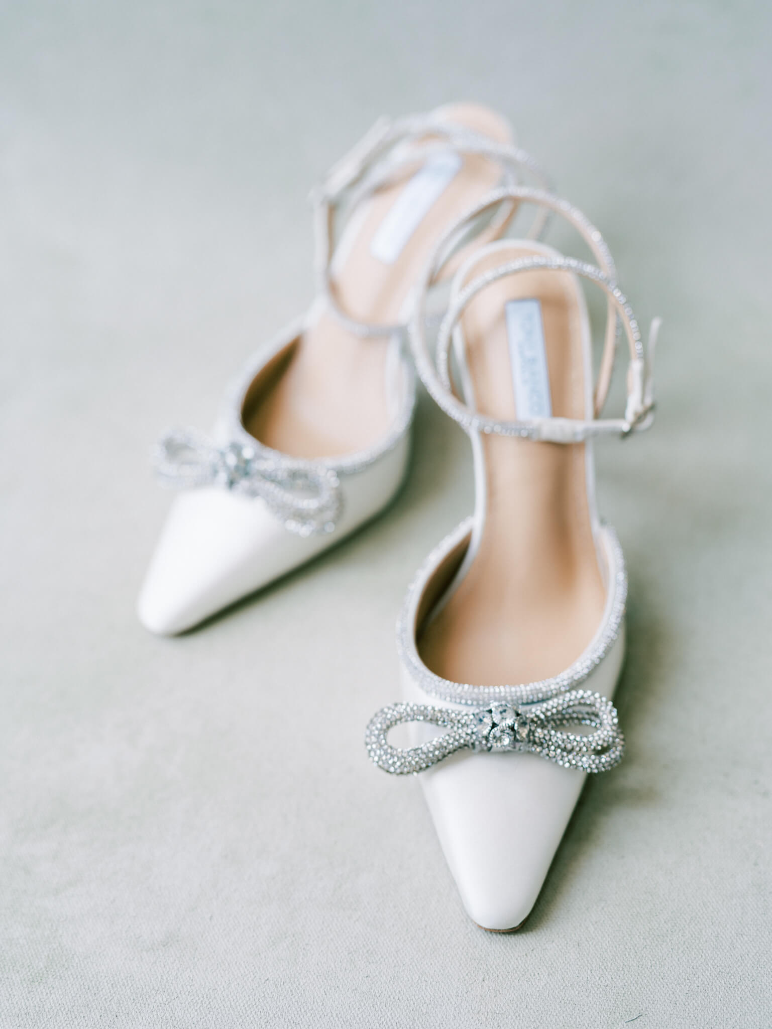 A pair of white wedding heels with rhinestone bows on a minty backdrop.