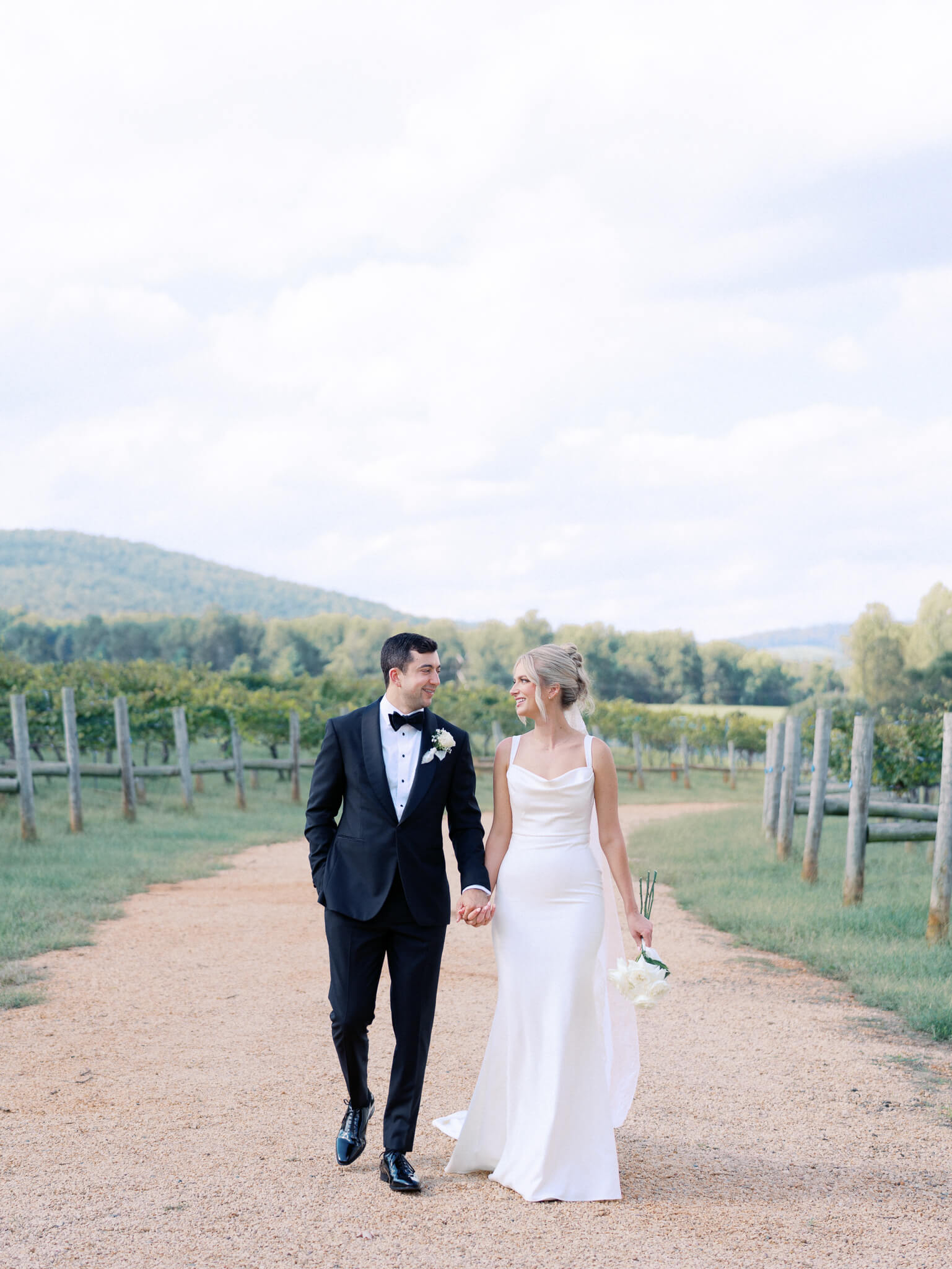 A bride and groom walking hand in hand on a dirt path through the grapevines, looking at each other.