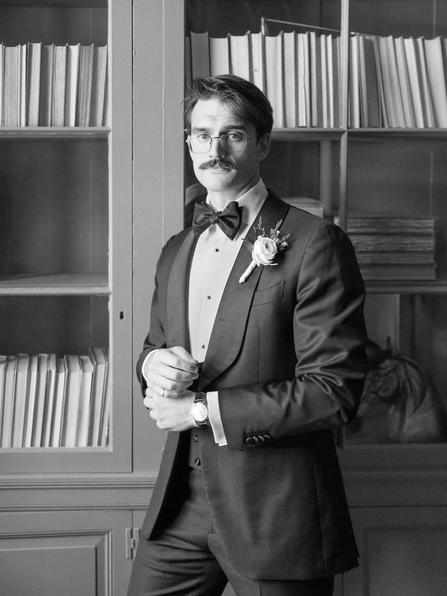 Closeup black and white image of a groom in a tux standing and leaning against a bookshelf in a library.