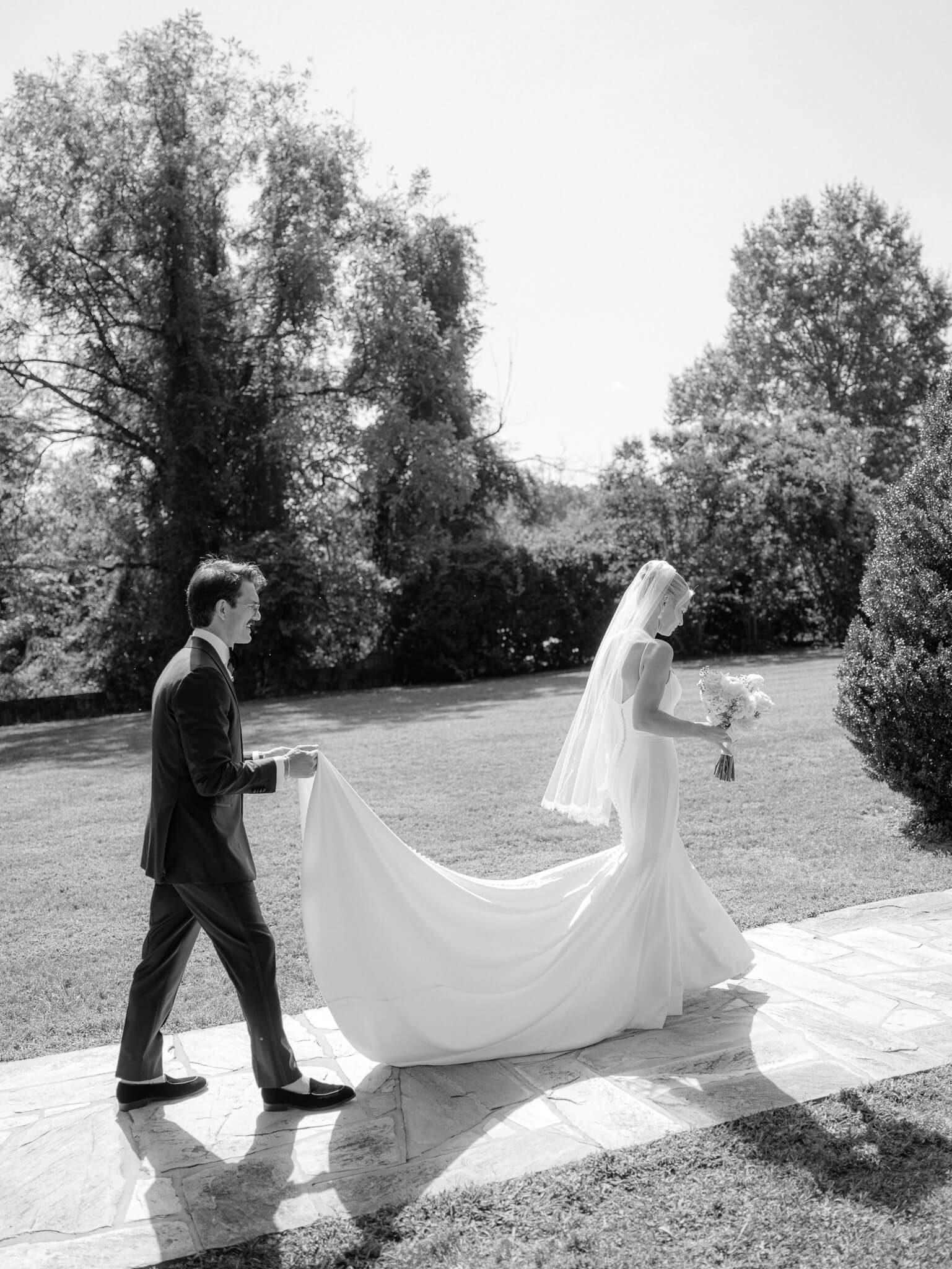 Black and white image of a bride walking ahead on a stone path while her groom in a tux is carrying the train of her wedding dress.