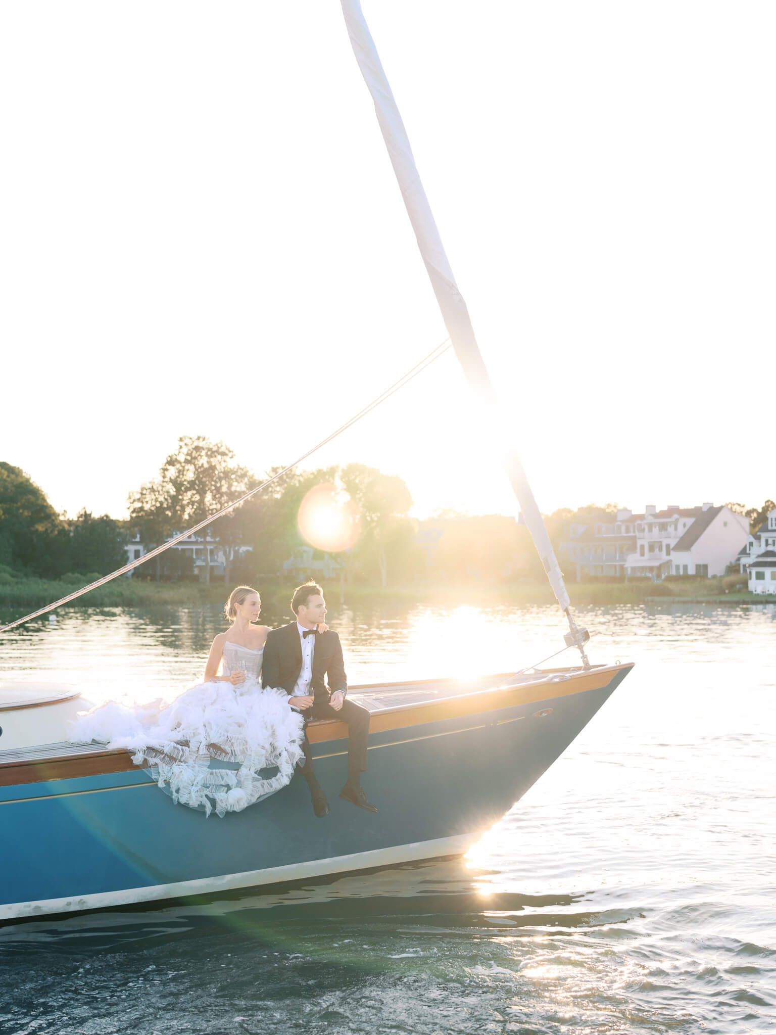 A bride and groom sitting on the front of a sailboat with the sunset and the Inn at Perry Cabin in the background.