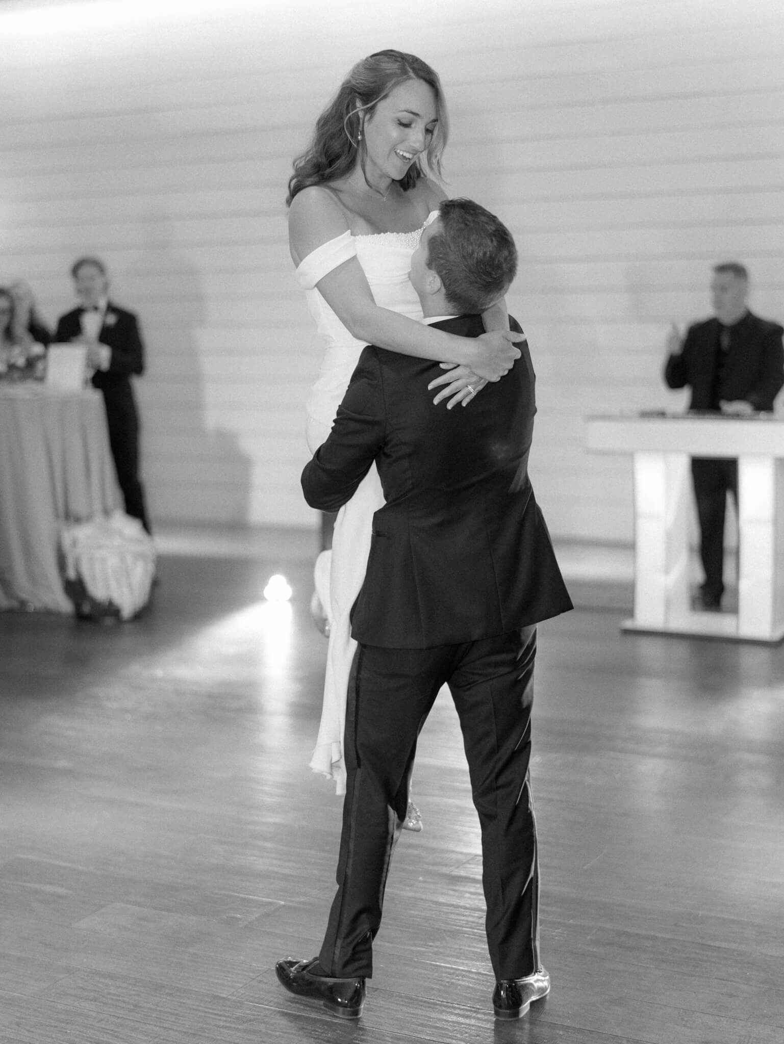 Black and white image of the groom picking up his bride during their first dance.