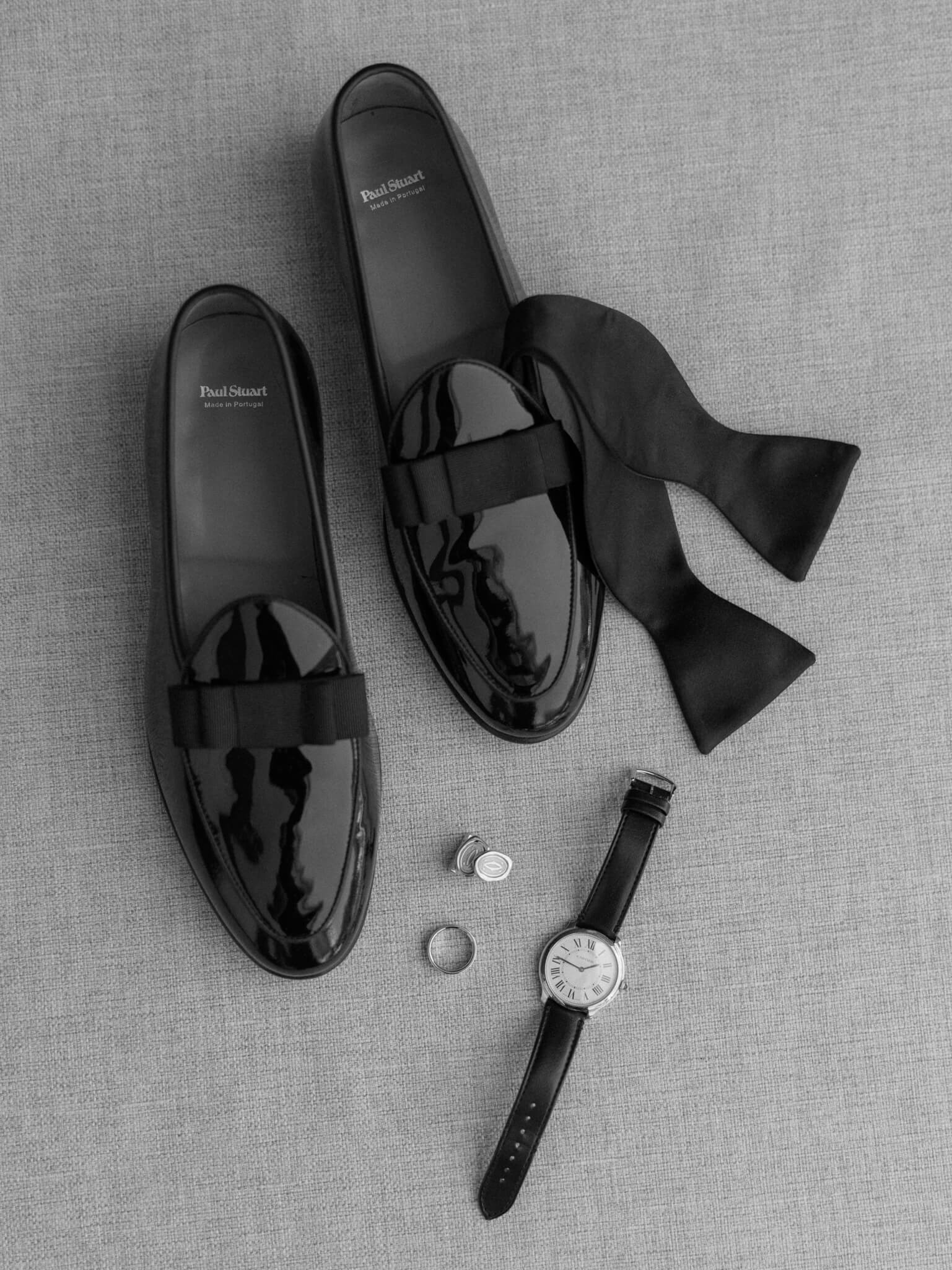 A black and white flatlay of a groom's shoes, his bowtie, watch, cufflinks, and wedding ring.