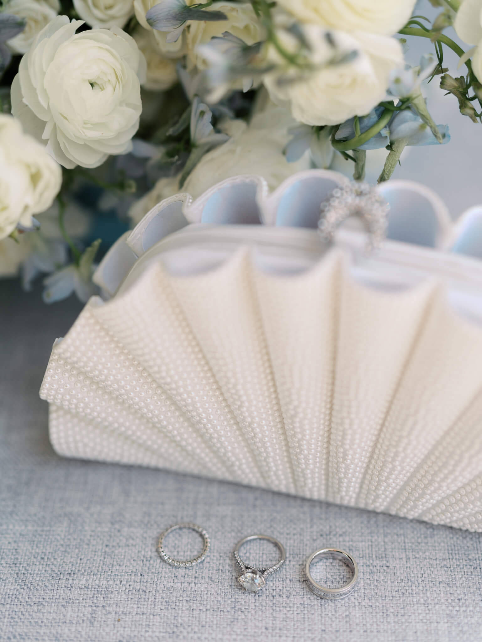 The wedding rings next to the bride's shell shaped clutch and her white and blue wedding flowers.
