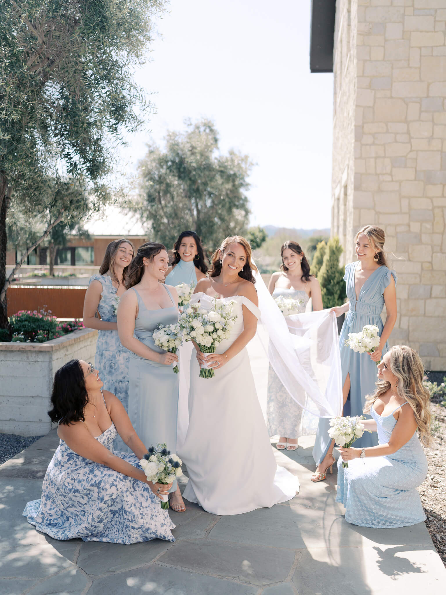 A bride surrounded by her bridesmaids in blue who are holding her veil.