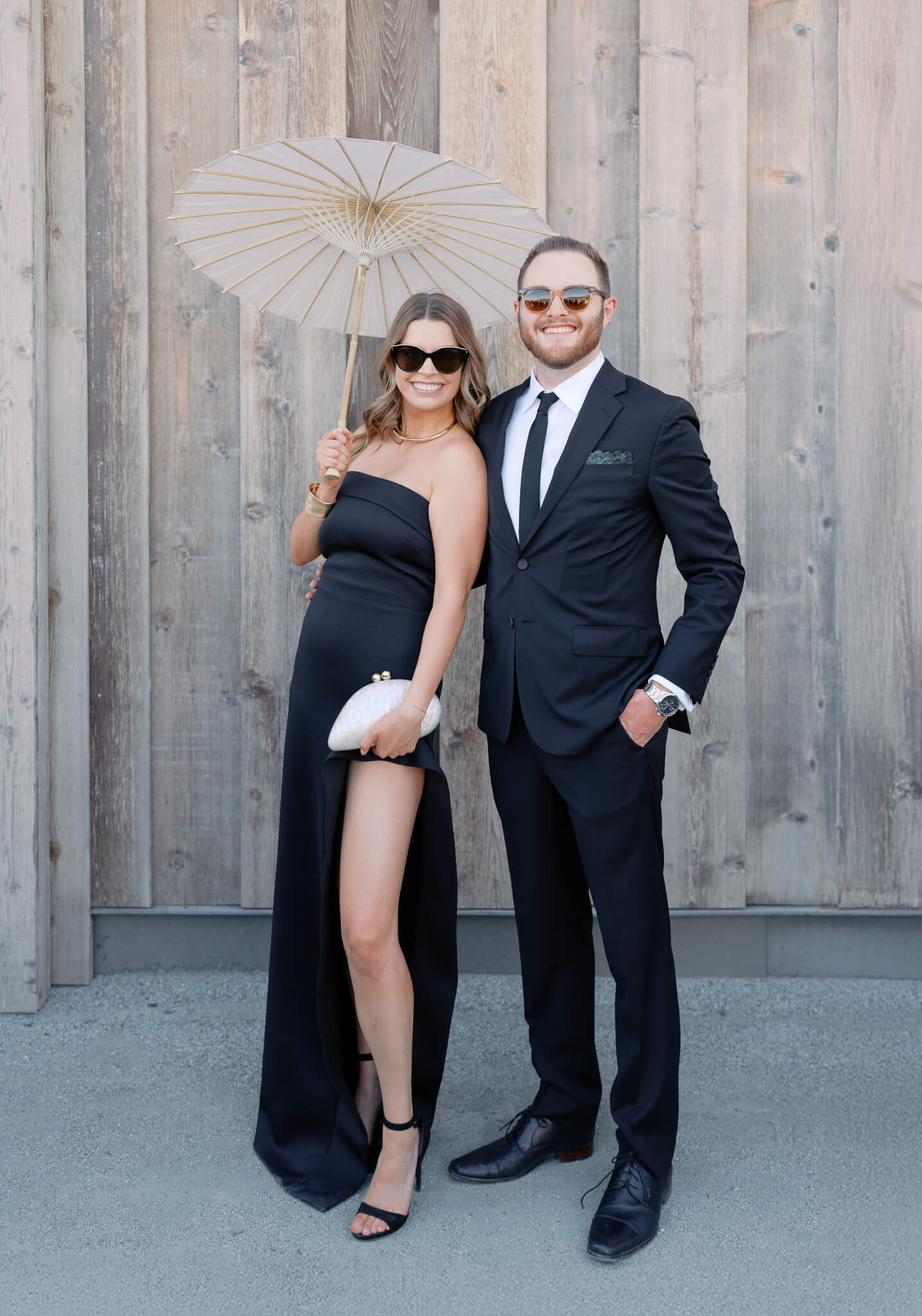 Wedding guests dressed in black, holding a parasol in front of a wooden barn backdrop.