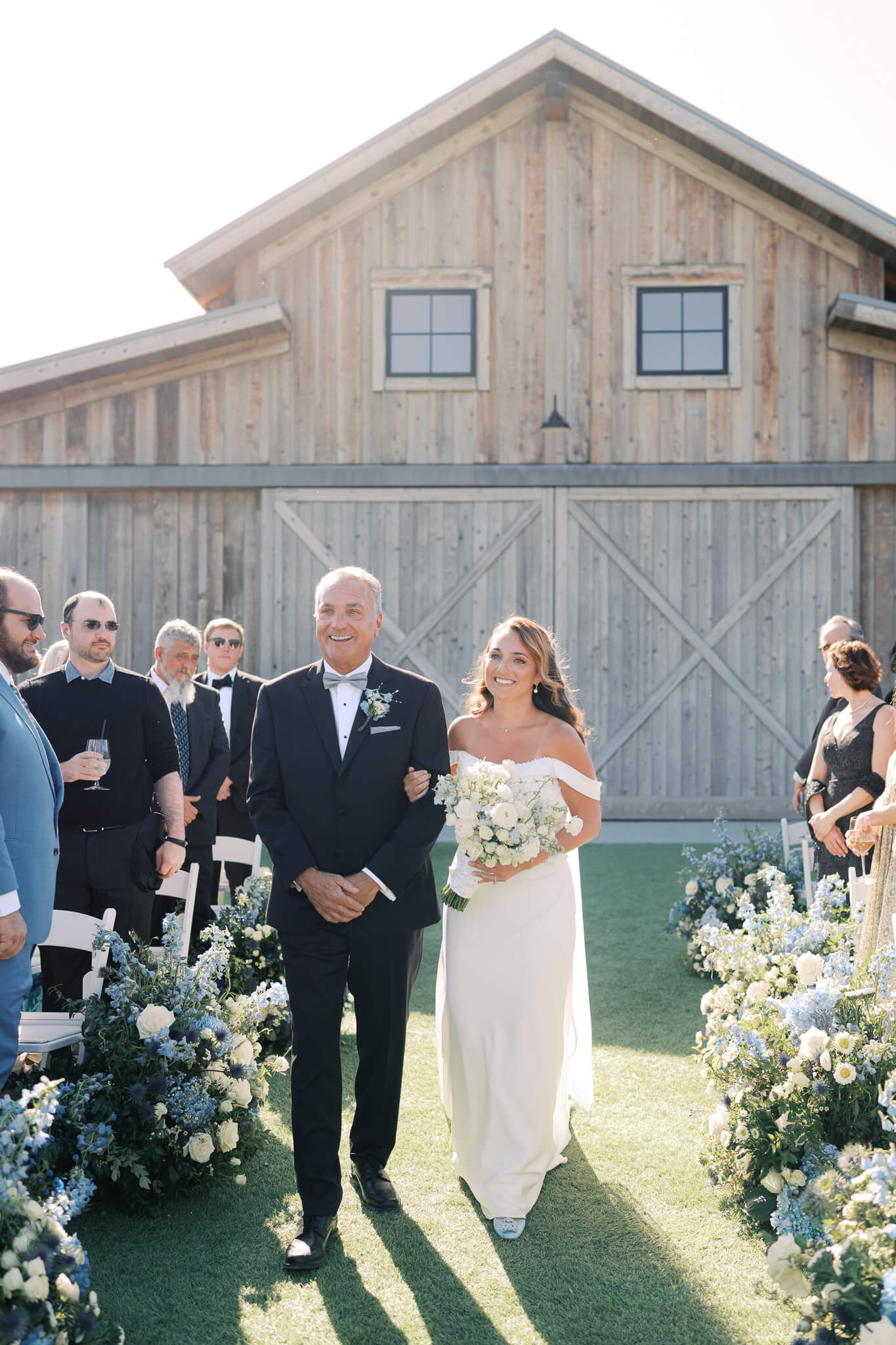 A bride and her father walking down the aisle in front of a barn building at a destination wedding weekend.