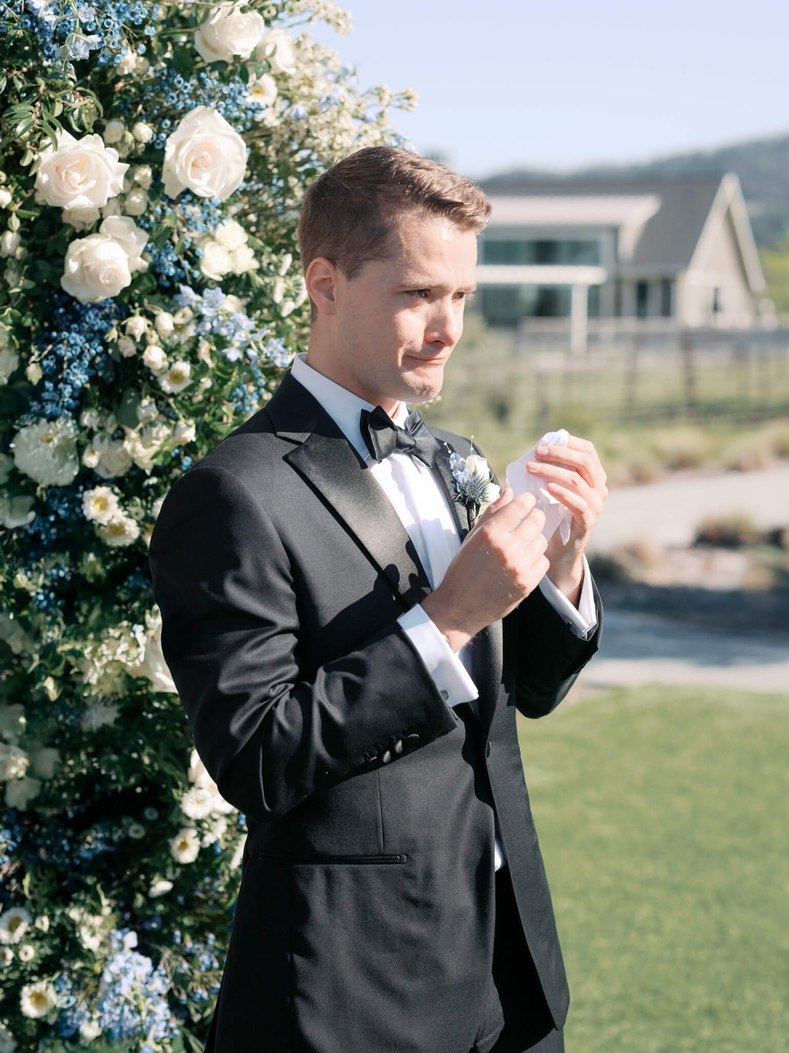 A groom dressed in a tux tearing up seeing his bride walk down the aisle.